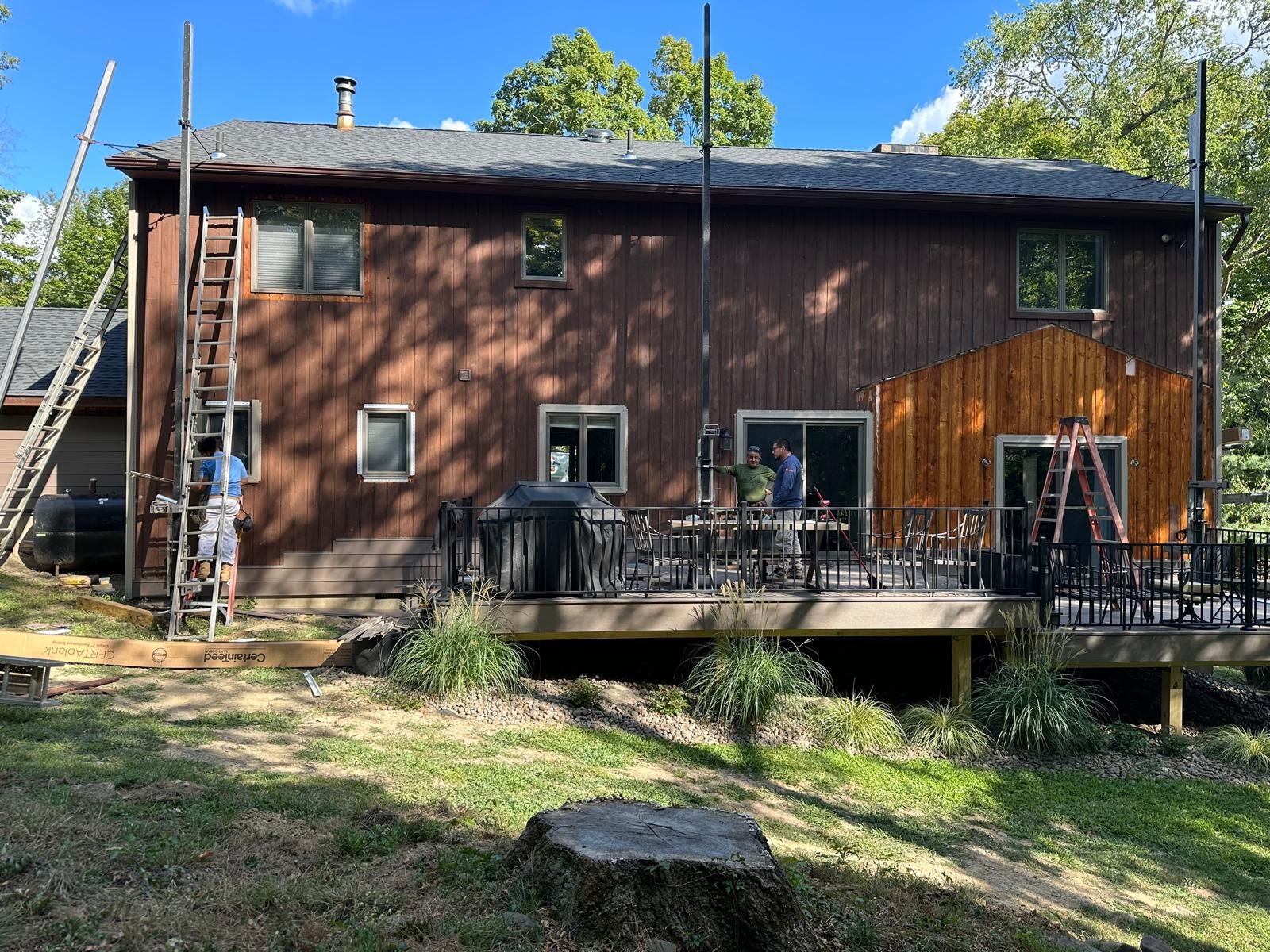 Two people work on the exterior of a brown two-story house with a wooden deck, ladders, and scaffolding.