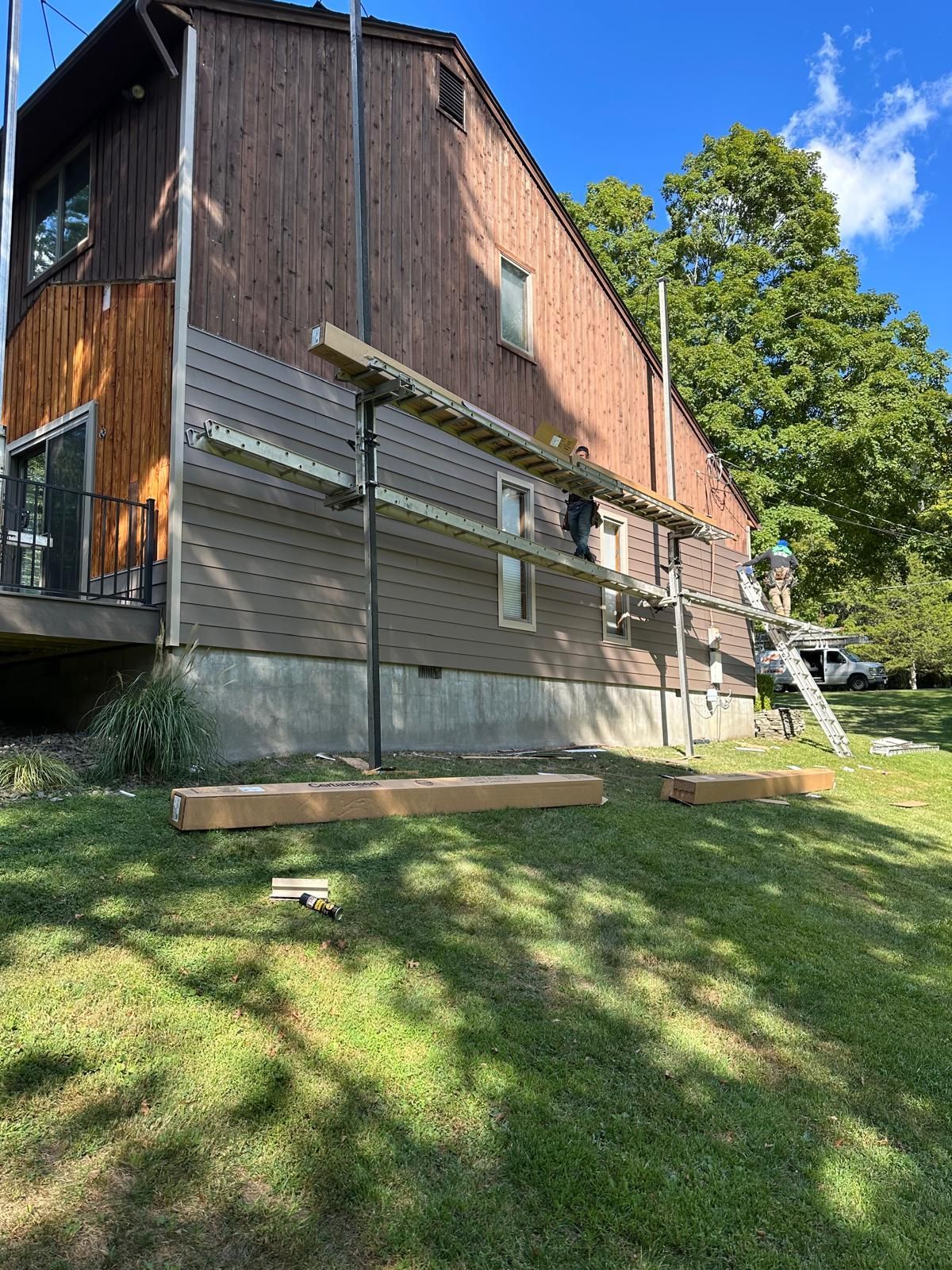 A person stands on scaffolding against the side of a house where new gray siding is being installed over wood paneling.