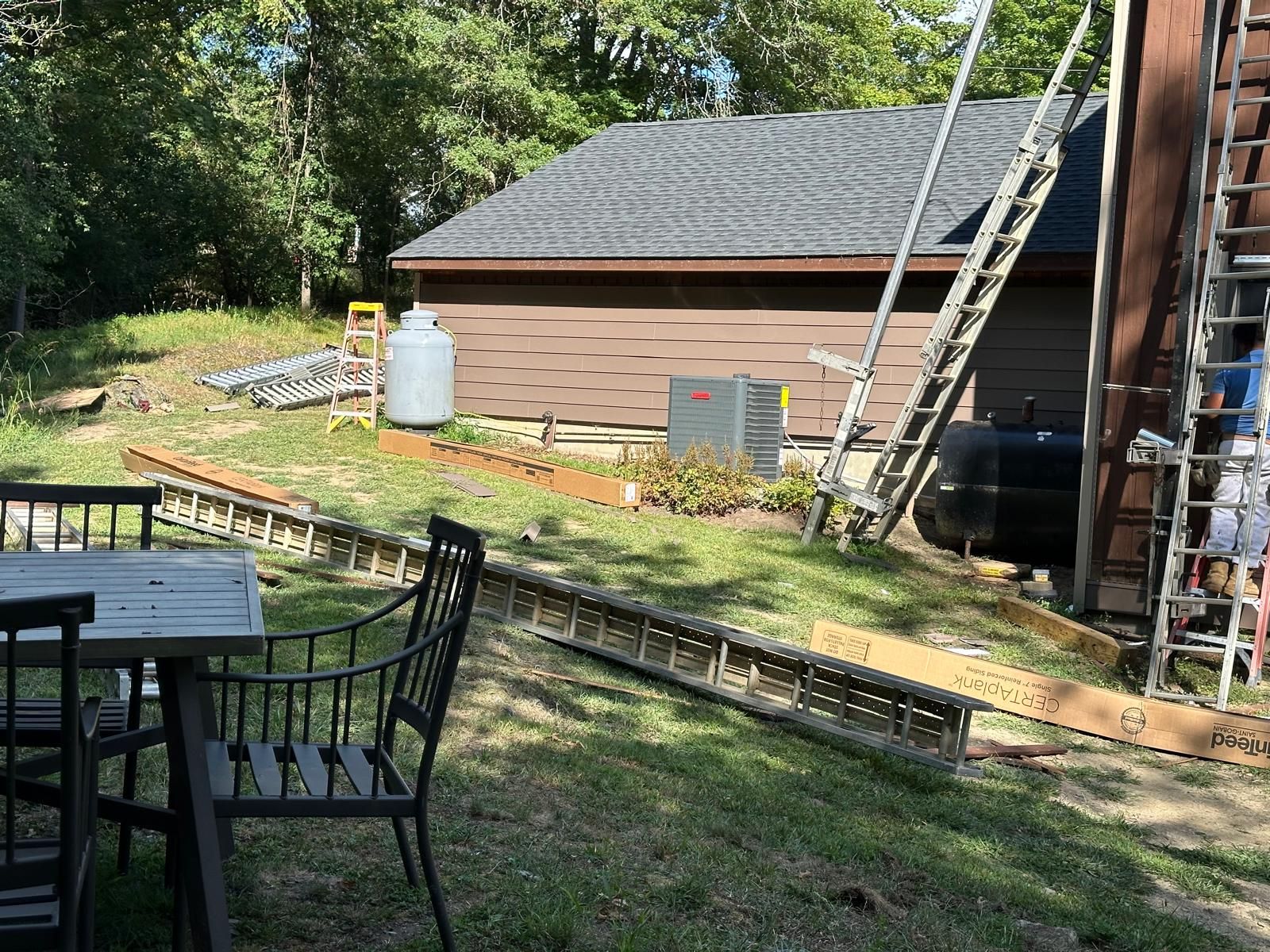 An outdoor scene featuring a patio table and chairs, with ladders, a shed, and equipment in the background.