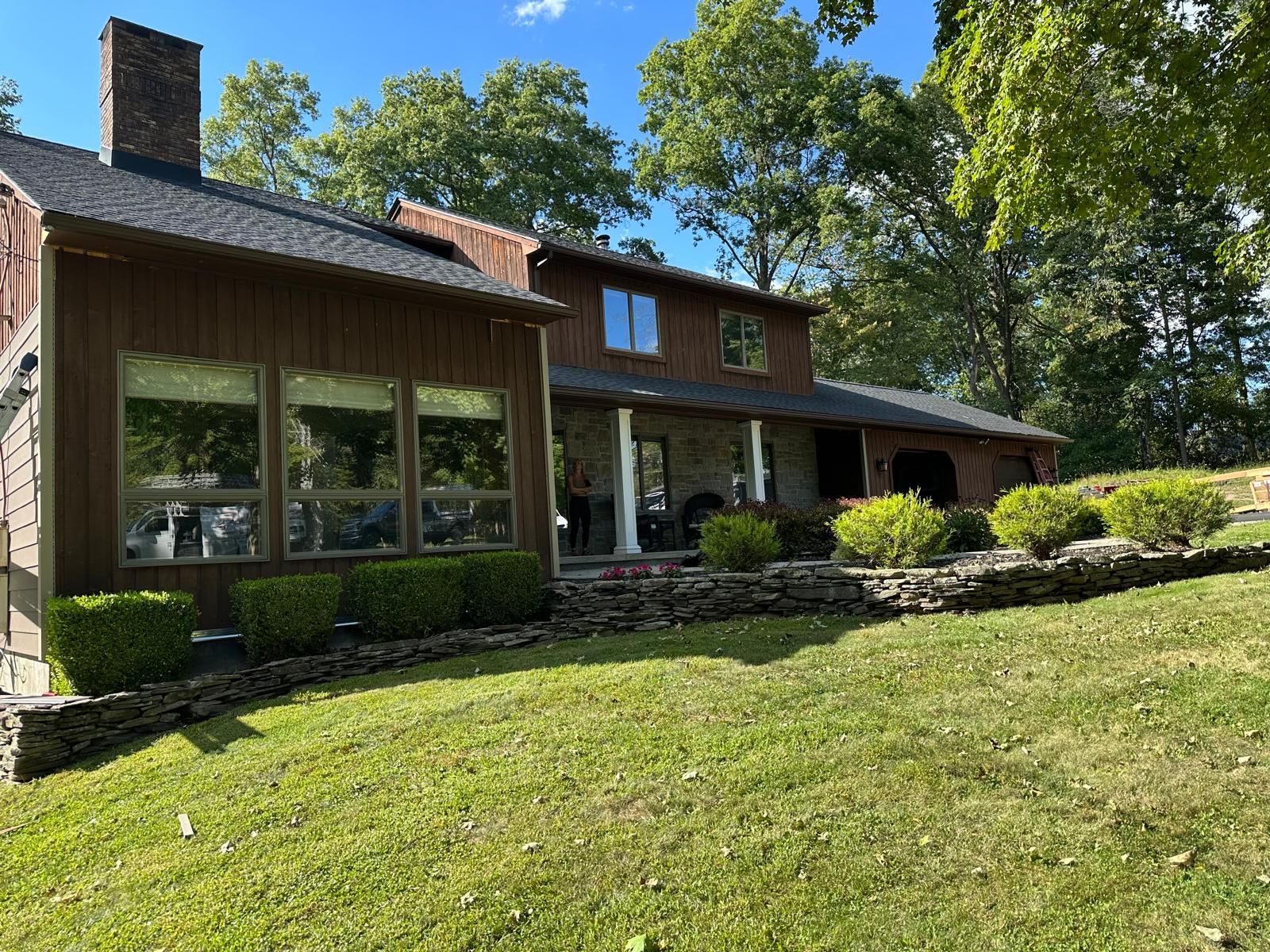 A two-story brown house with large windows and stone landscaping, nestled among green trees on a sunny day.