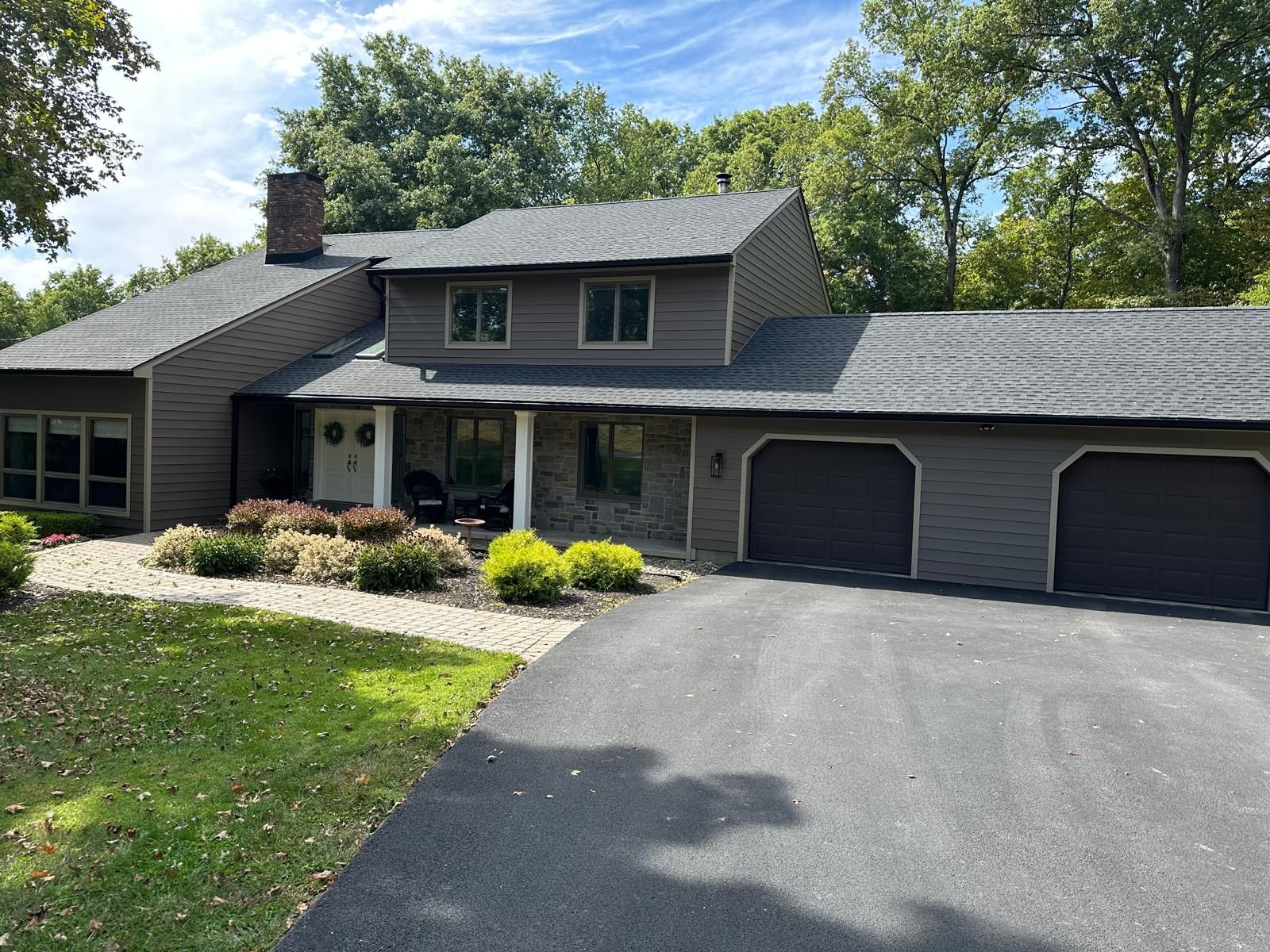 A two-story brown house with a gray shingled roof, two garage doors, and a stone-accented front porch.