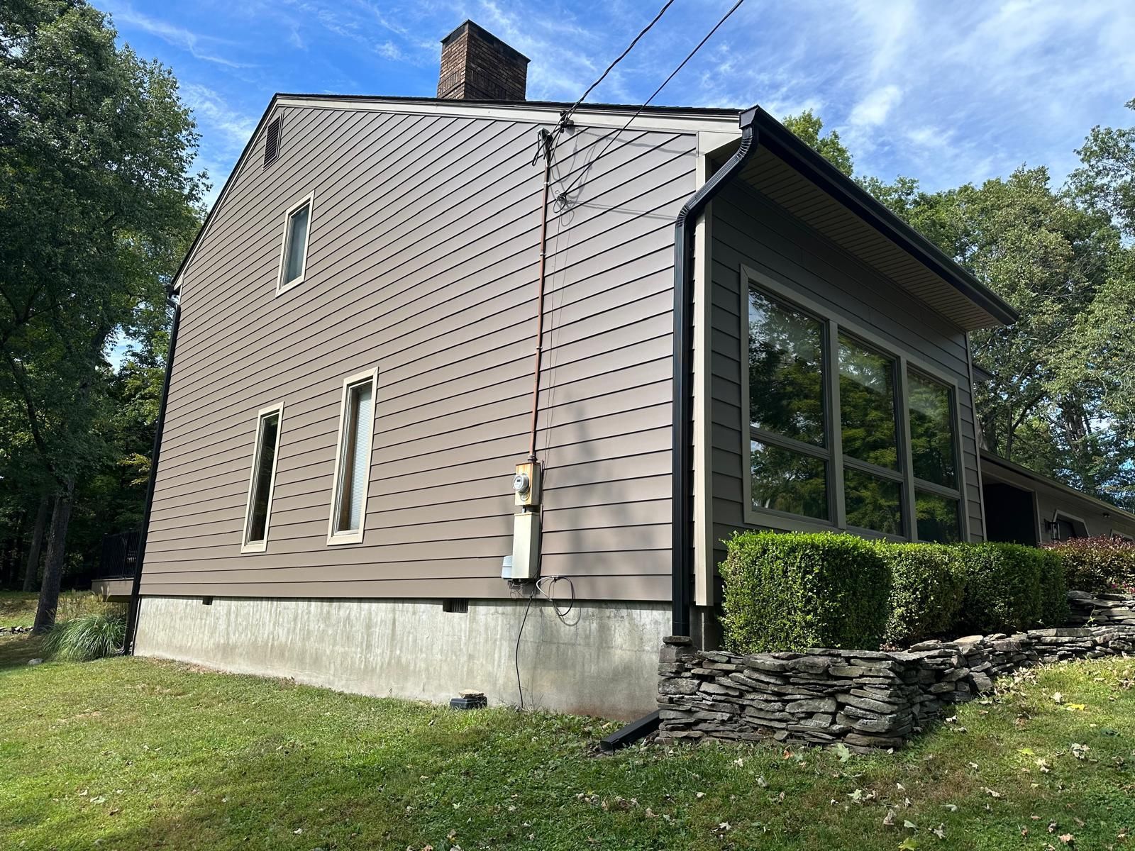 A side view of a brown two-story house with horizontal siding, a concrete foundation, and large windows set on a lawn.