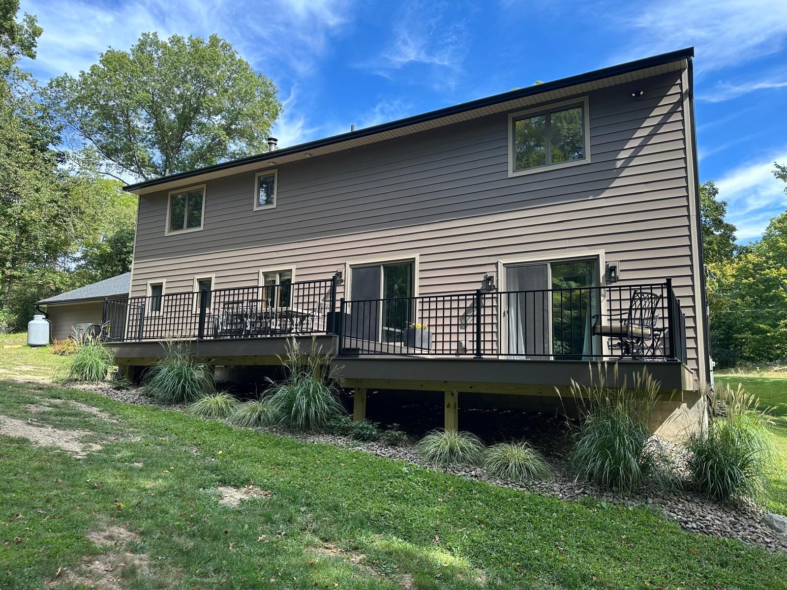 A two-story tan house with horizontal siding and a black-railed deck, set against a grassy yard under a blue sky.