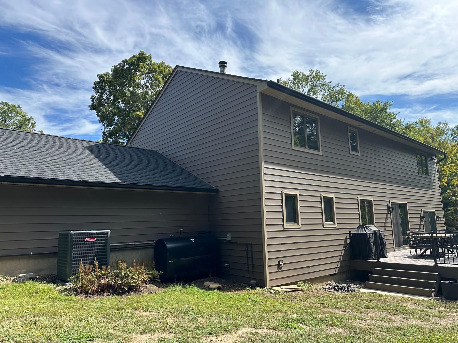 The side view of a brown, two-story house with horizontal siding, a gray shingled roof, a back deck, and yard equipment.