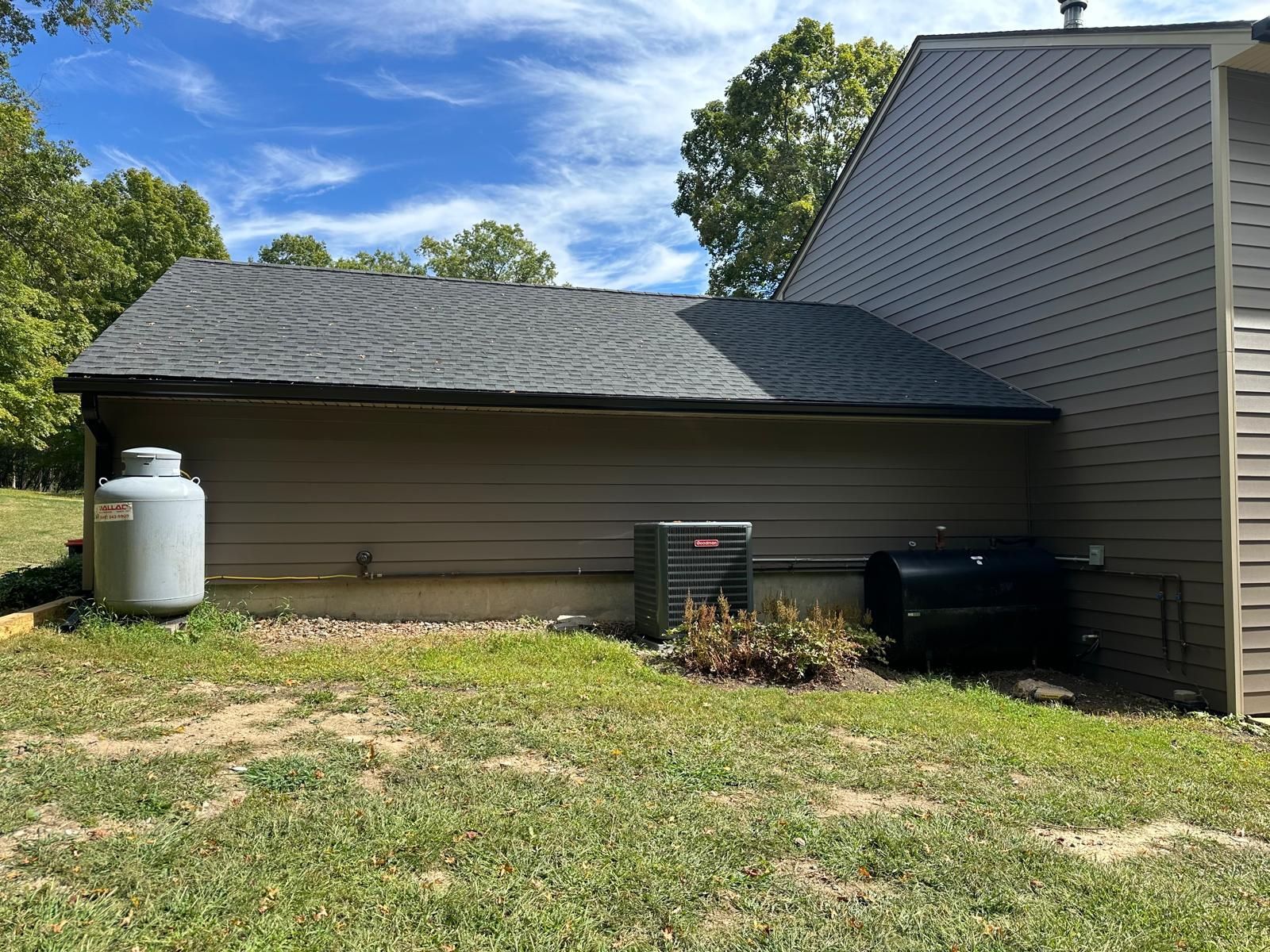 The side of a brown-sided house with a dark shingle roof, a grey propane tank, an AC unit, and a black storage bin.