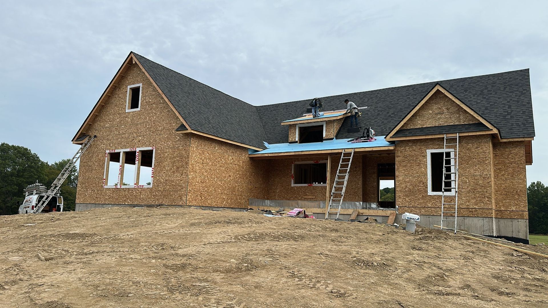 A house under construction with exposed wooden framing and a partially shingled roof on a dirt lot.