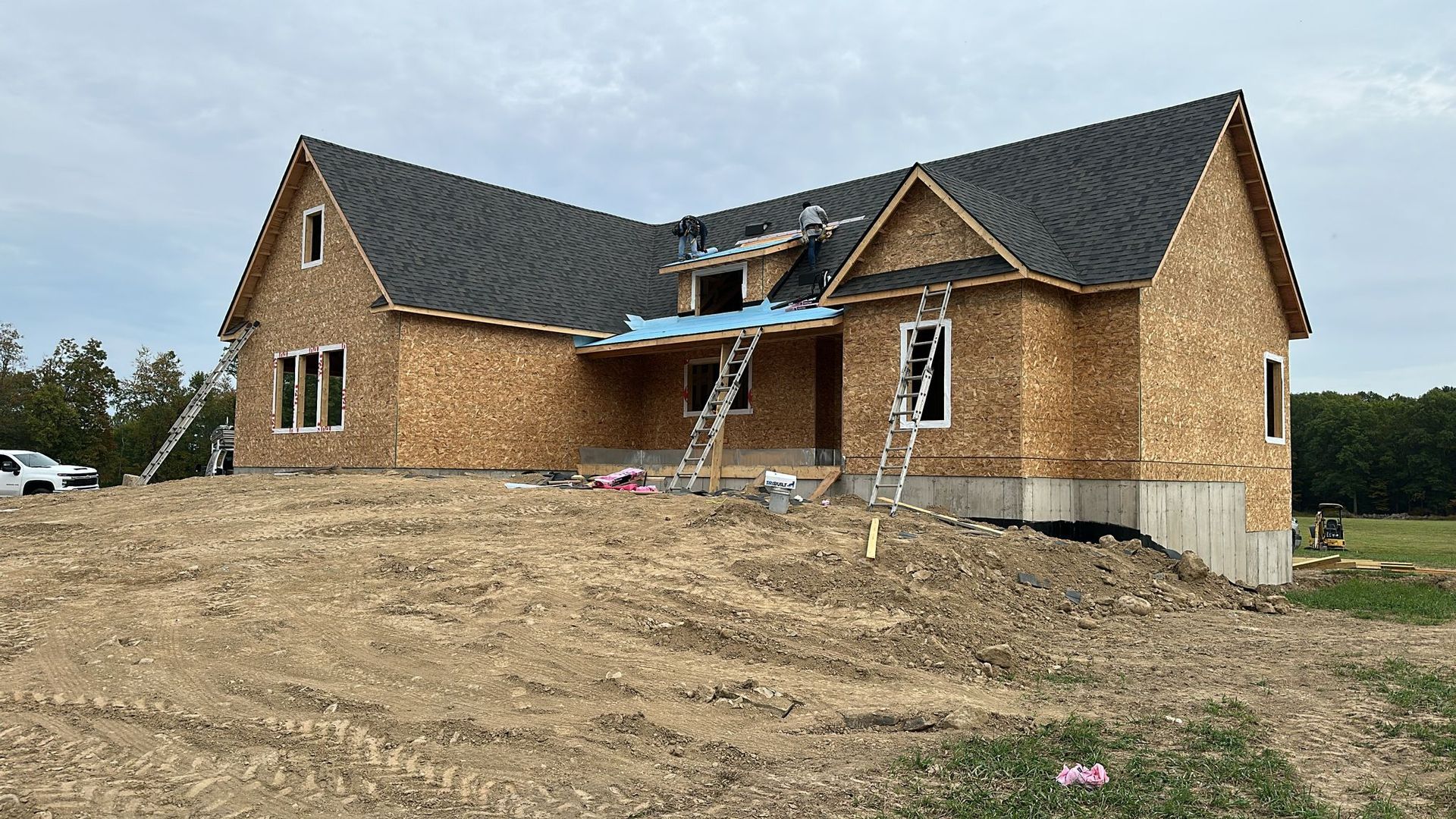 A house under construction with a dark shingled roof and wood-paneled walls on a dirt lot under a cloudy sky.