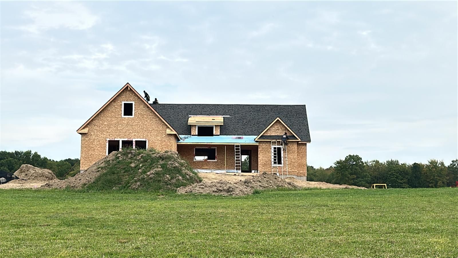 A house under construction with a dark roof and wooden framing, situated in a wide, grassy field under a cloudy sky.