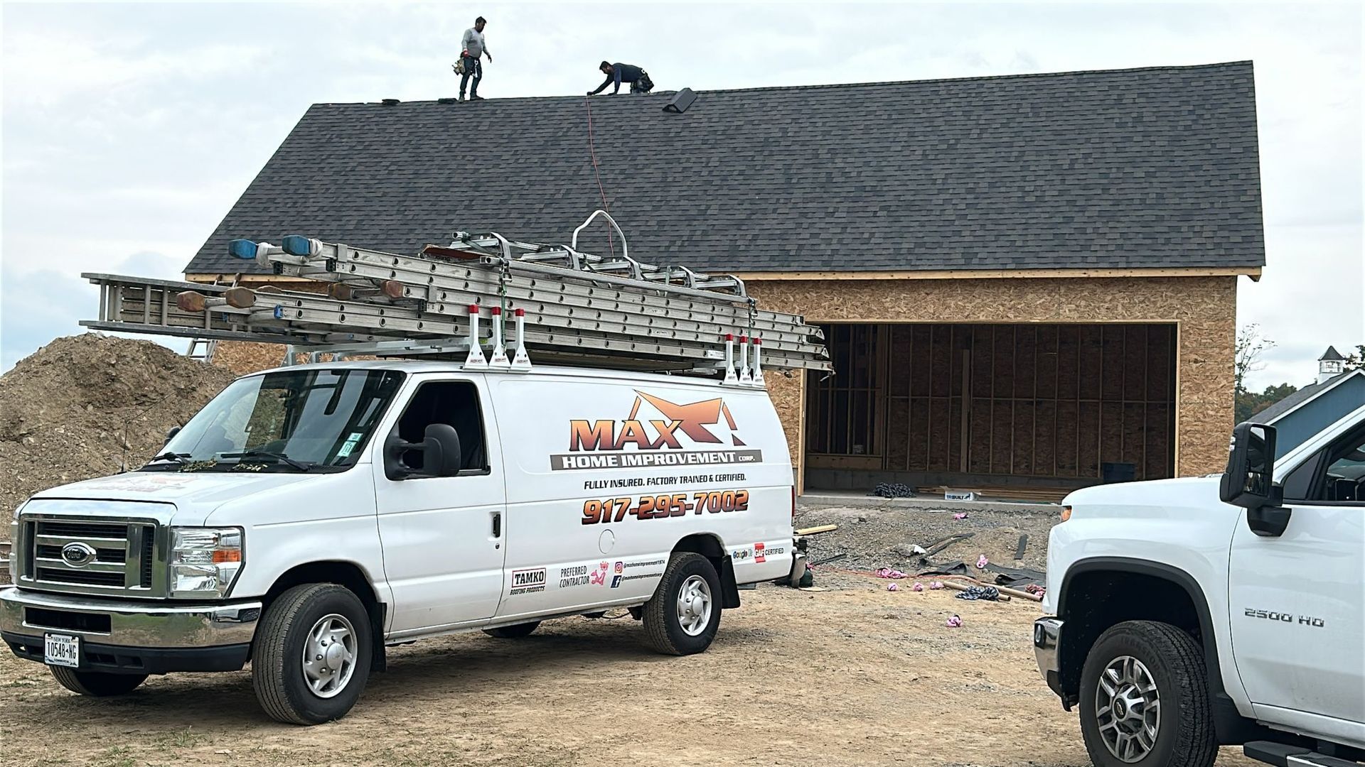 Two workers install shingles on the roof of a house under construction, with a company van parked in the foreground.