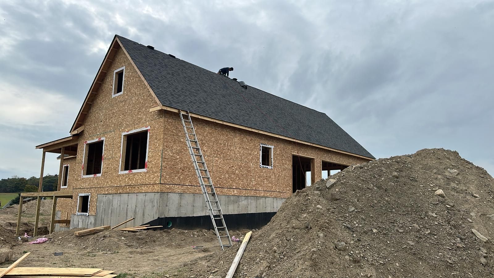 A construction site with a partially finished house featuring dark shingles and OSB walls, next to a large pile of dirt.