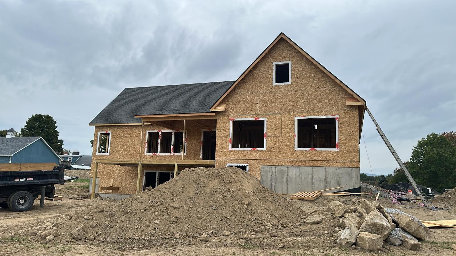 A two-story house under construction with exposed wooden frame, dark shingled roof, and a dirt mound in the front yard.