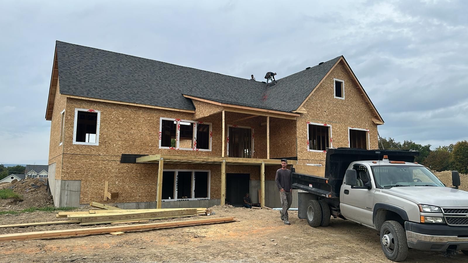 A partially built wooden house frame with a dark shingled roof, a front porch, and a dump truck parked in the foreground.
