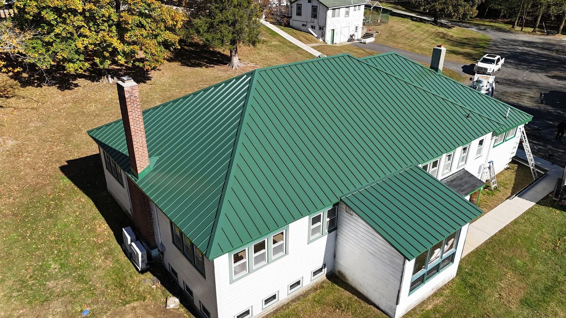 A high-angle view of a white house with a green standing-seam metal roof, a brick chimney, and a gravel driveway.