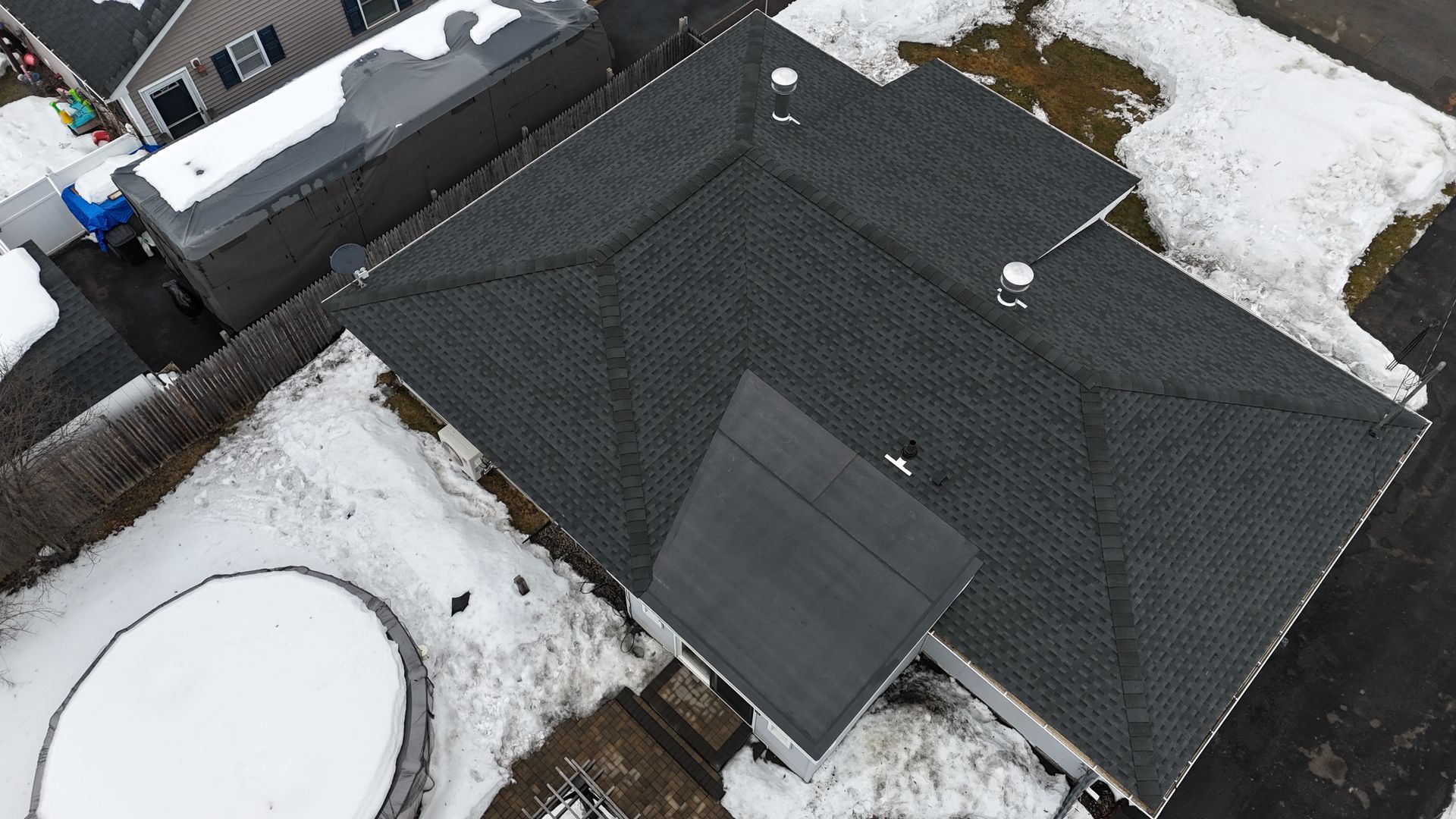 Aerial view of a dark shingled roof with snow patches and a small dormer in a residential area