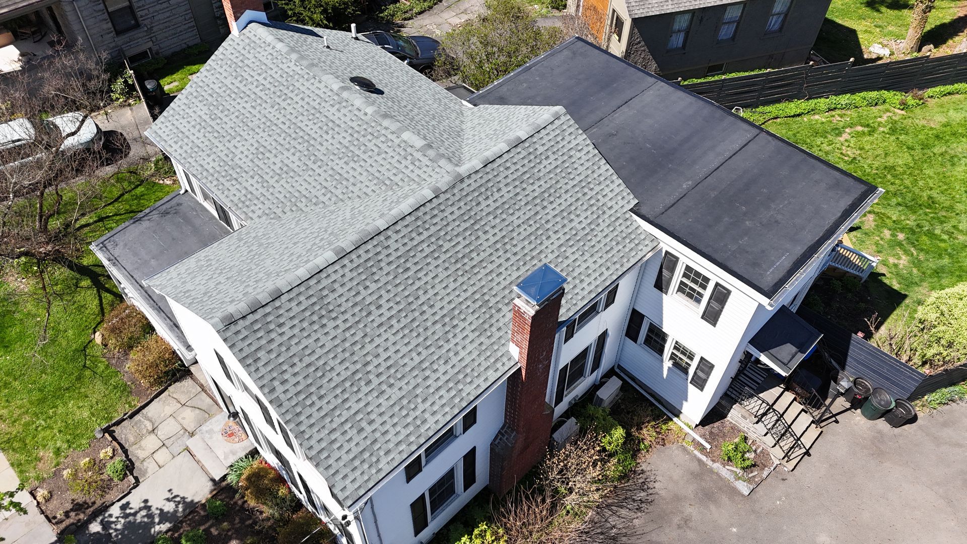 Aerial view of a white house with gray roofs, a brick chimney, and a driveway surrounded by greenery.