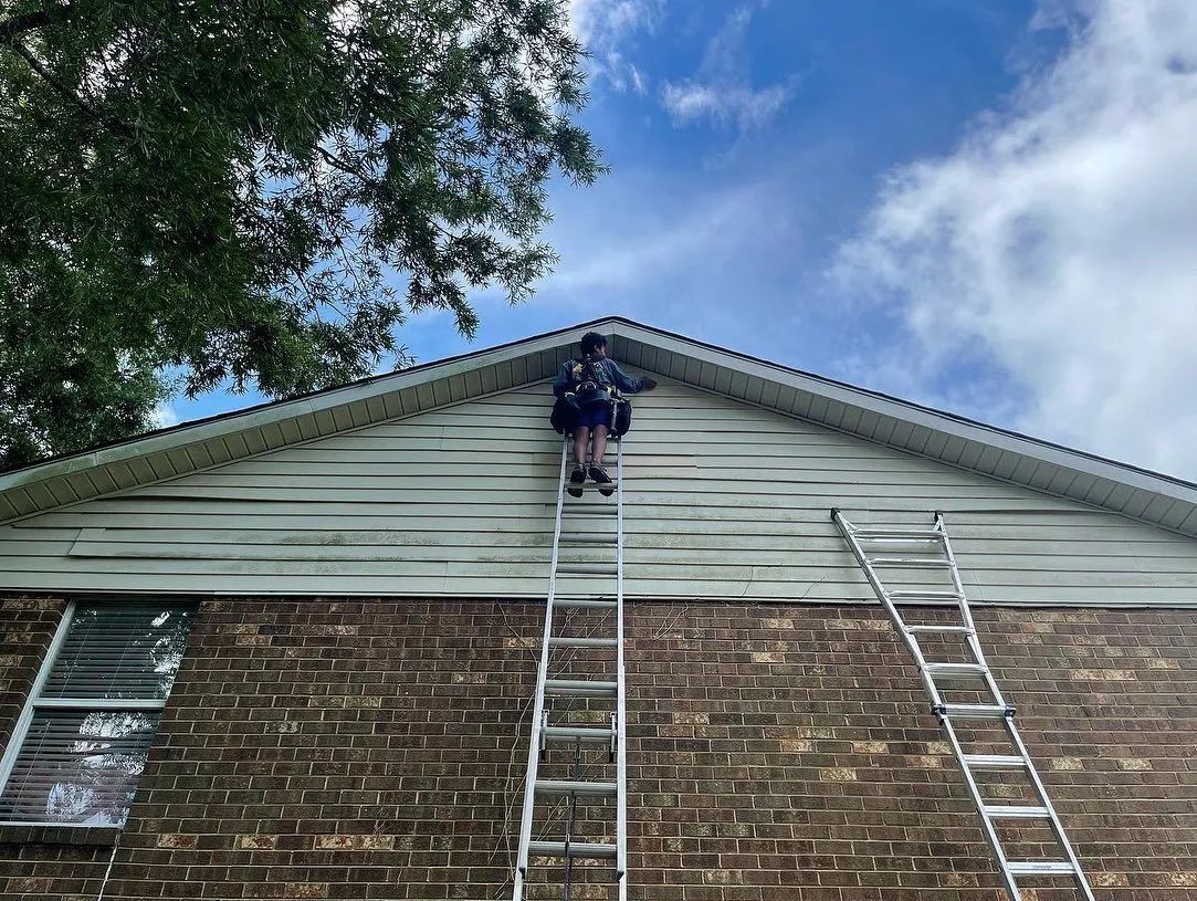 Person on a ladder working on siding on a house under a cloudy sky.