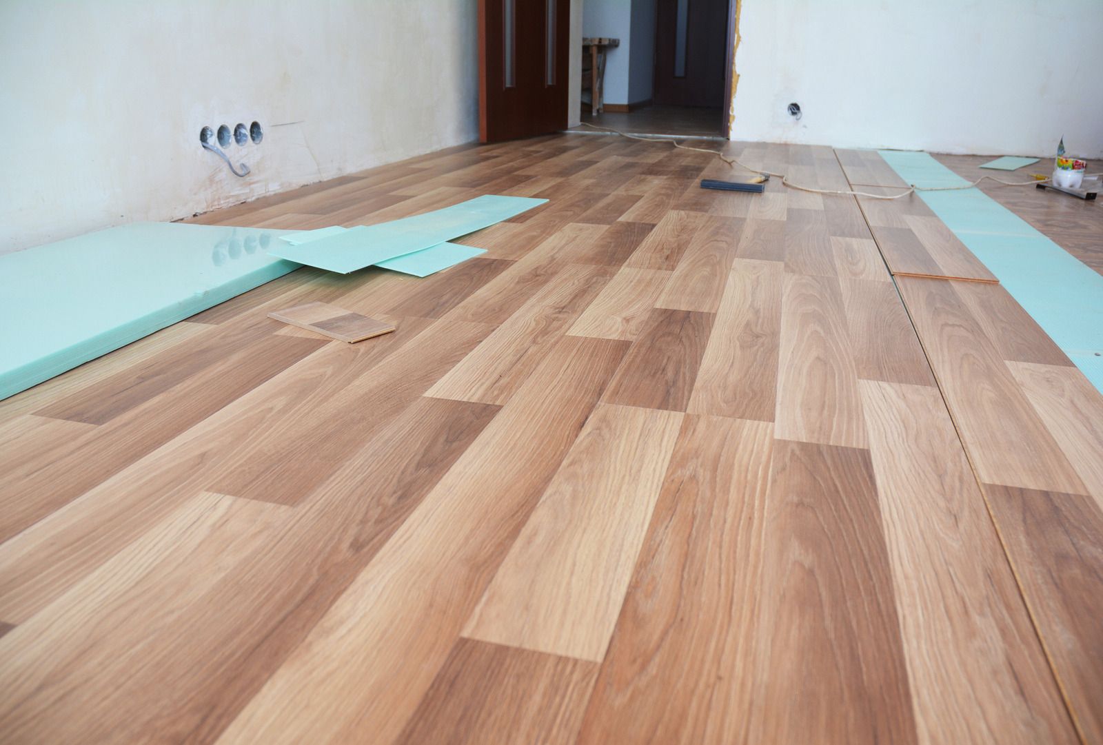 Wood-look laminate flooring being installed in an empty room, with blue underlayment sheets along the walls.