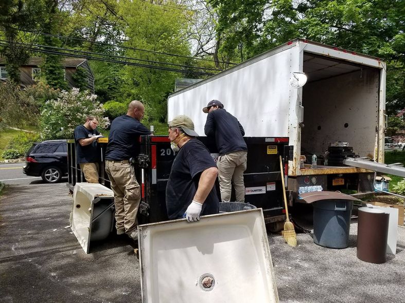 Four workers load a shower base and other items into a large moving truck parked on a gravel driveway.