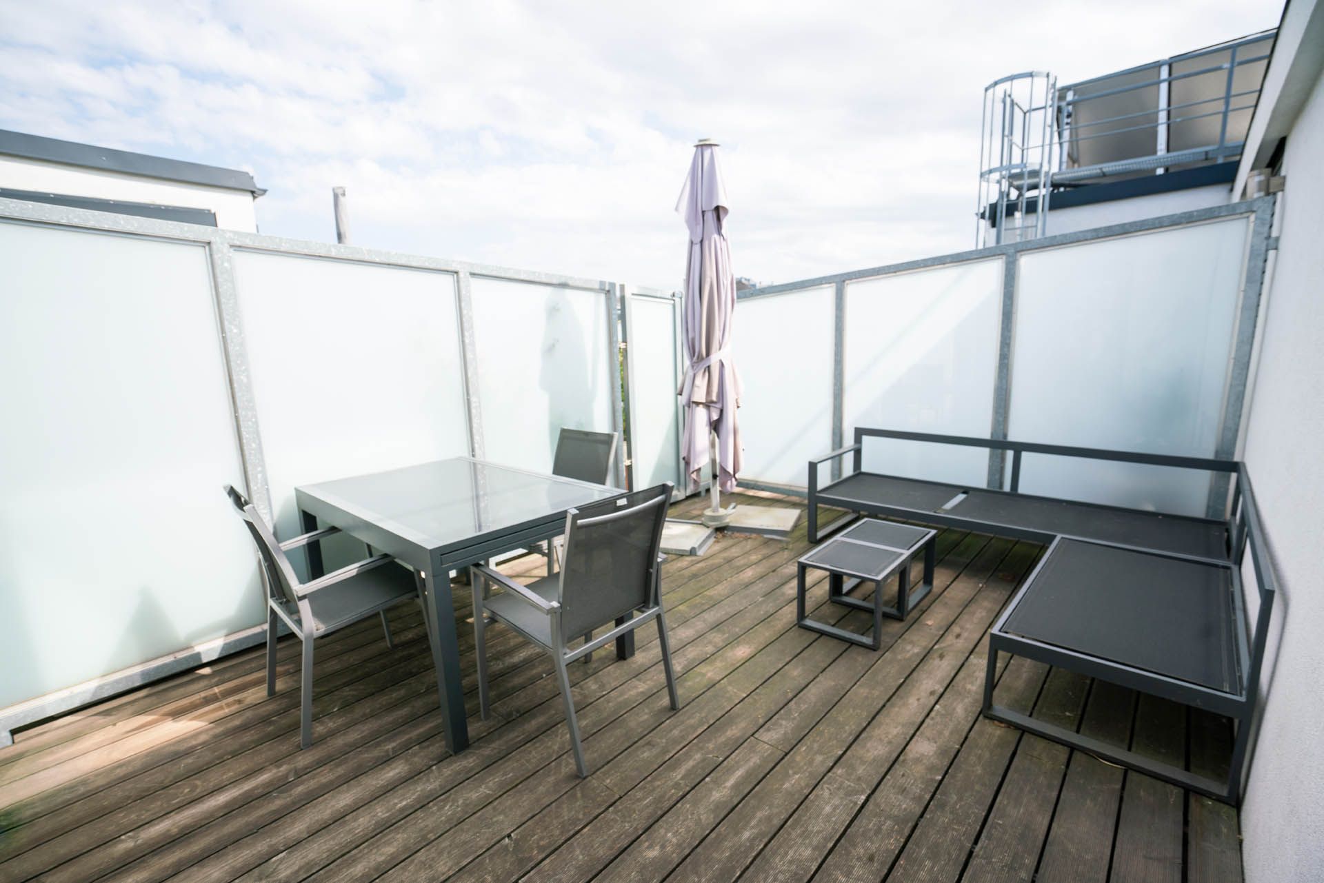 Terrasse mit Tisch, Stühlen, Sofa und teilweise zusammengeklapptem Sonnenschirm, umgeben von Milchglasplatten.