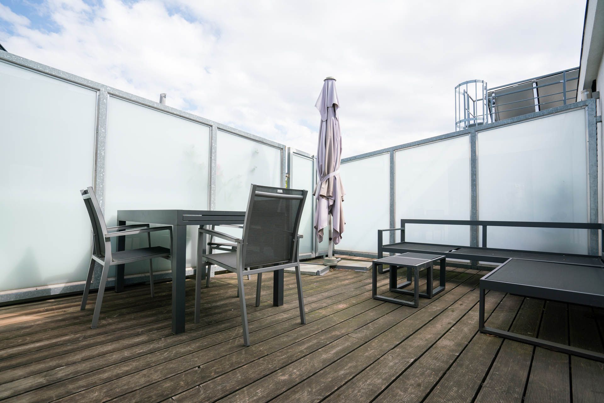 Terrasse mit grauen Möbeln, einem geschlossenen Sonnenschirm und durchscheinenden Wänden vor einem bewölkten Himmel.