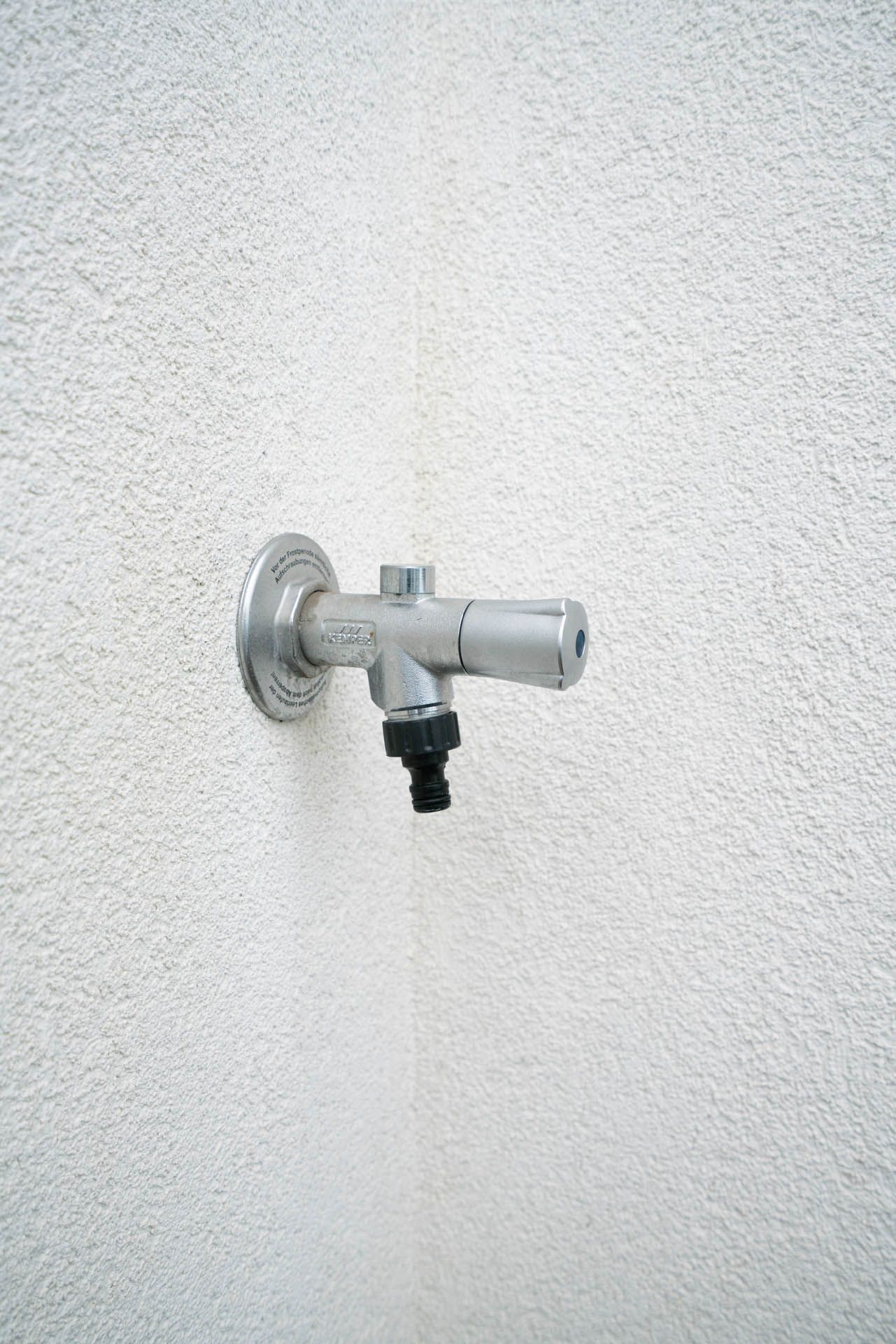 Silberner Außenwasserhahn an einer strukturierten weißen Wand, mit einem schwarzen Schlauchanschluss.