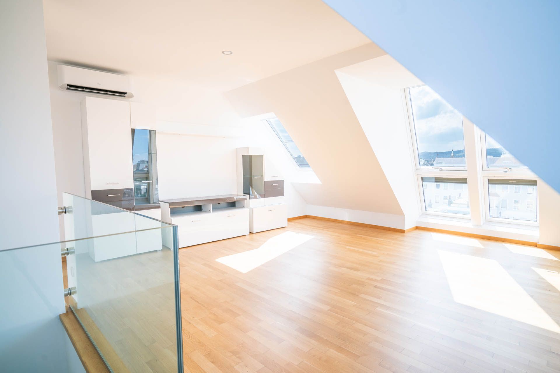 Heller, leerer Wohnraum mit schräger Decke, Oberlicht und großem Fenster. Holzböden und weiße Schränke.