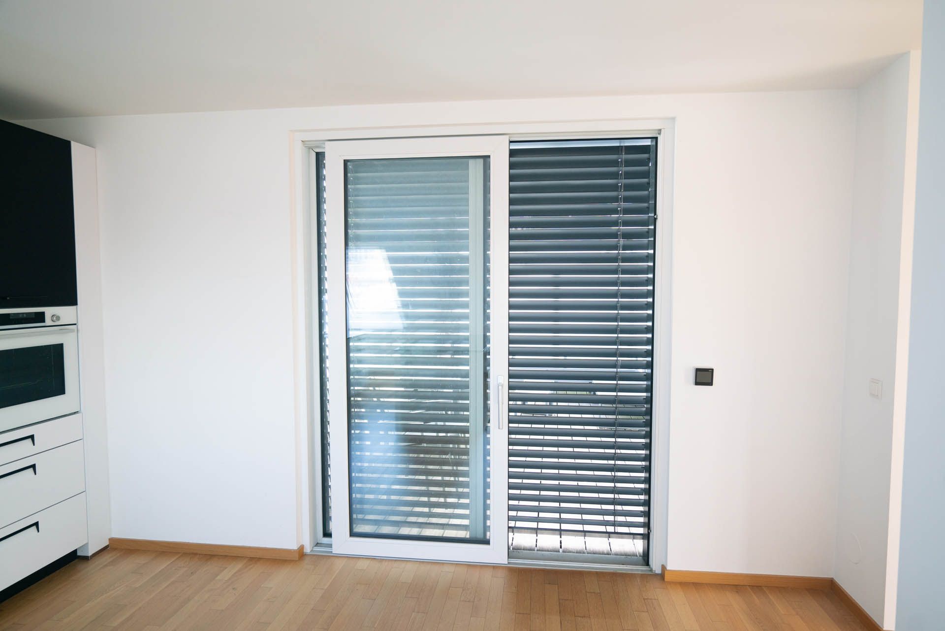 Raum mit weißen Wänden und einer Glasschiebetür mit Jalousien. Holzboden und ein weißer Schrank sind sichtbar.