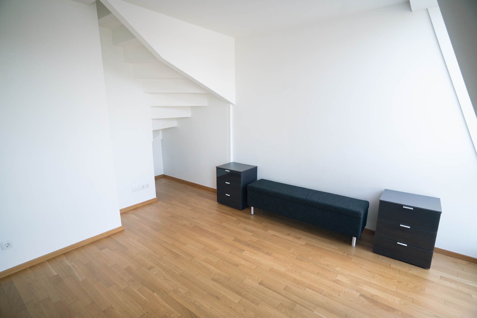 Leerer Raum mit Holzboden, weißen Wänden, schwarzen Möbeln und einer Treppe, die nach oben führt.