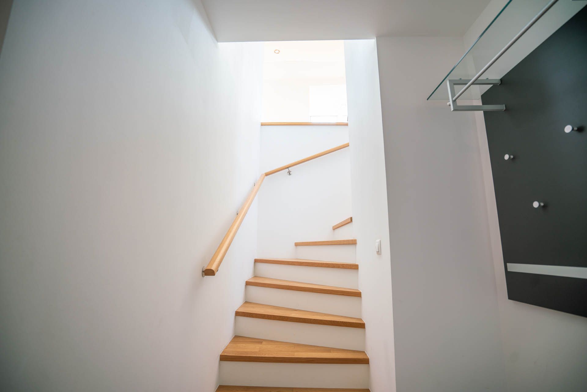 Treppe mit Holzstufen und Handlauf, die zu einer Öffnung hinaufführt. Zu sehen sind weiße Wände und eine Garderobe.