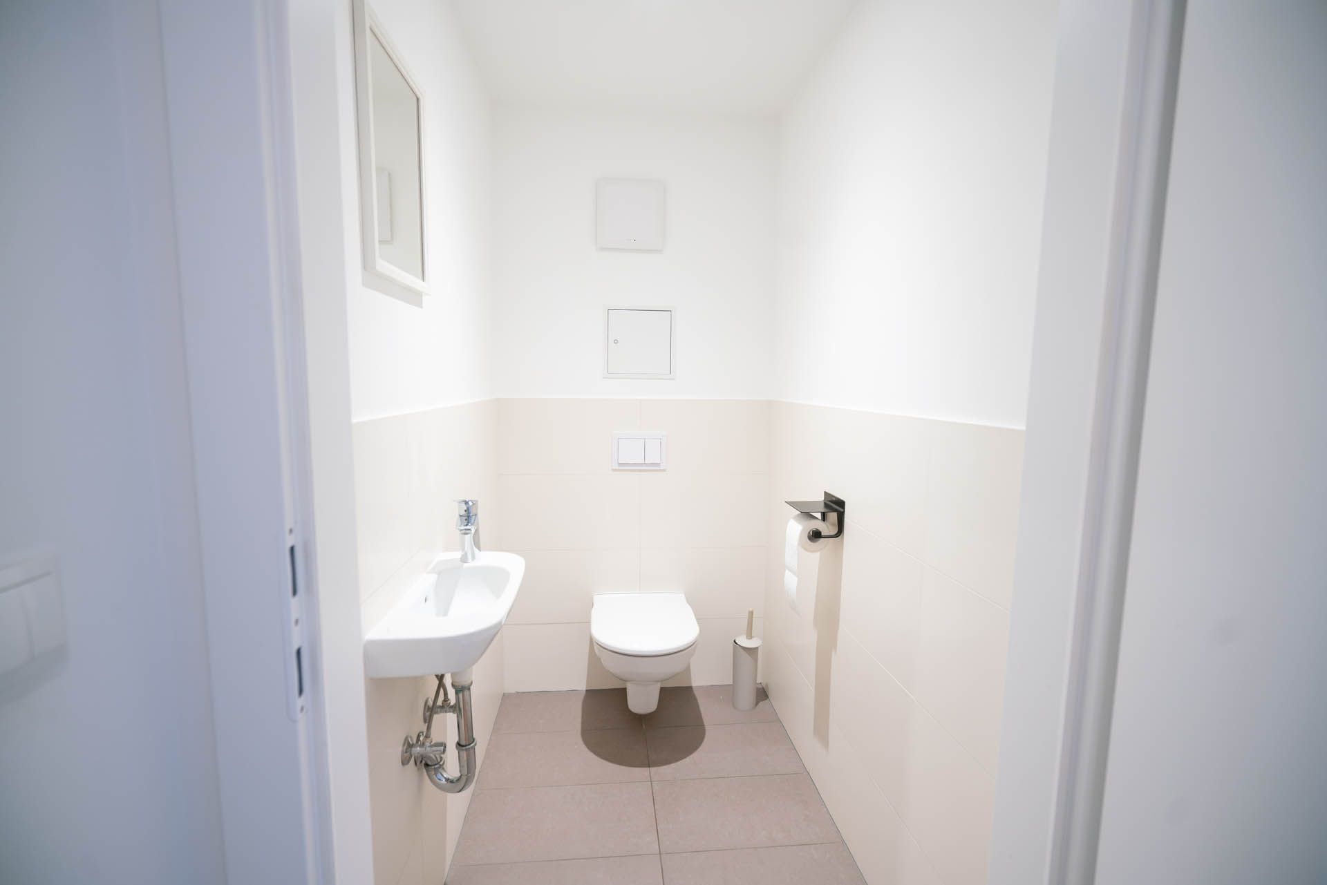 Kleines, weißes Badezimmer mit Toilette, Waschbecken und offener Tür. Grauer Fliesenboden.