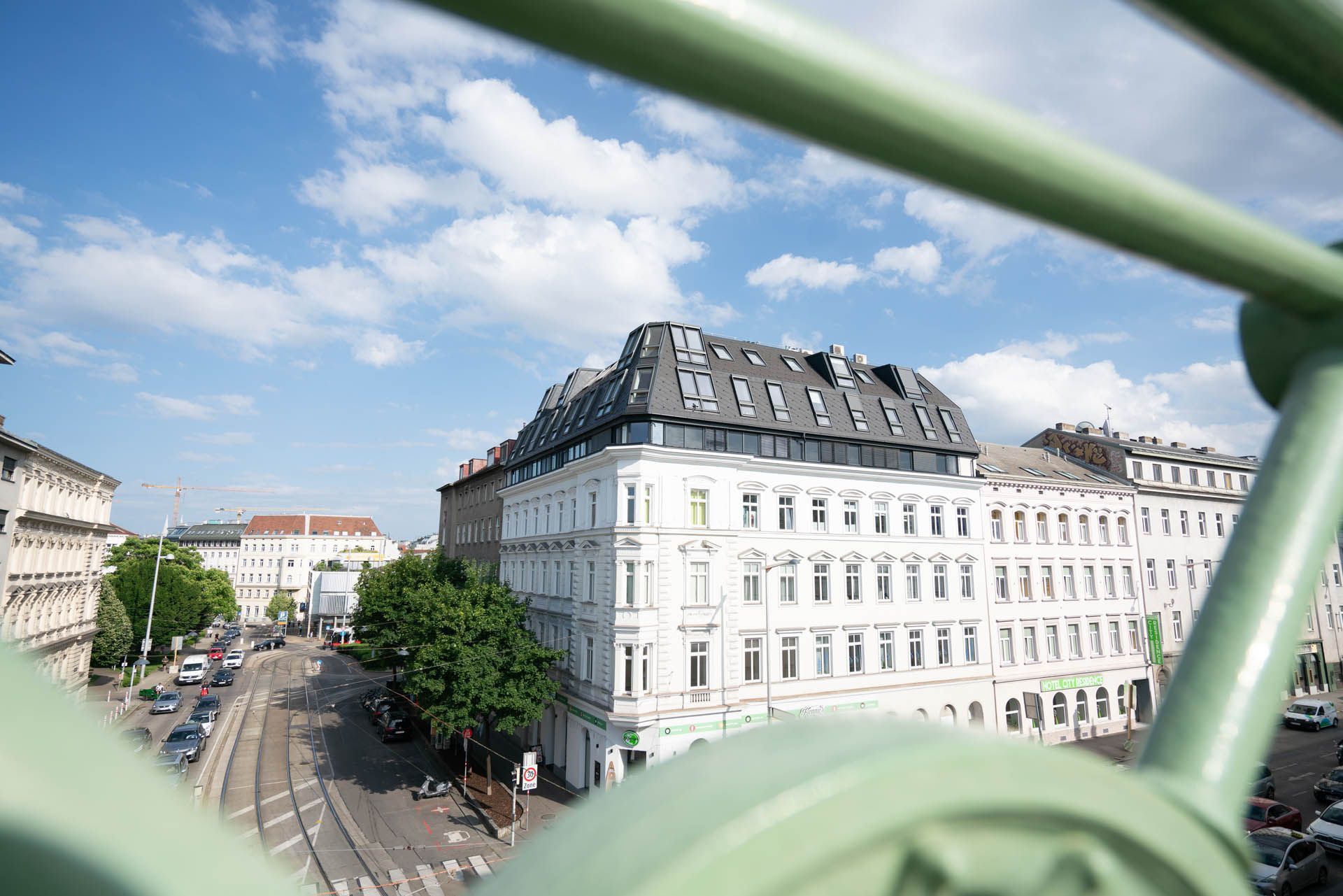 Blick auf ein weißes Gebäude mit grauem Dach durch einen grünen Metallrahmen, unter einem blauen Himmel mit Wolken.