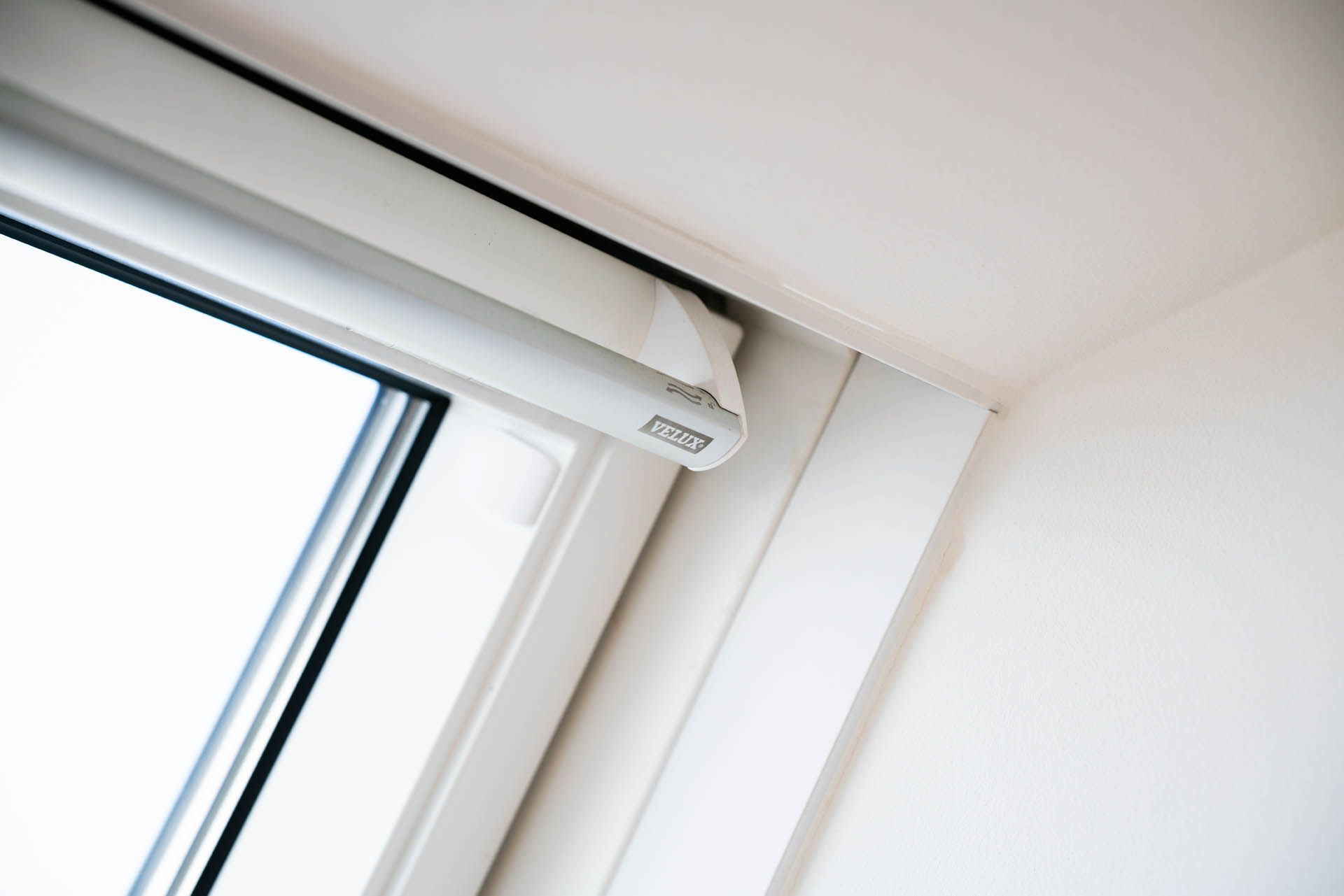 Weißer Fensterrahmen mit einer teilweise hochgerollten weißen Jalousie. Die obere rechte Ecke des Raumes ist sichtbar.