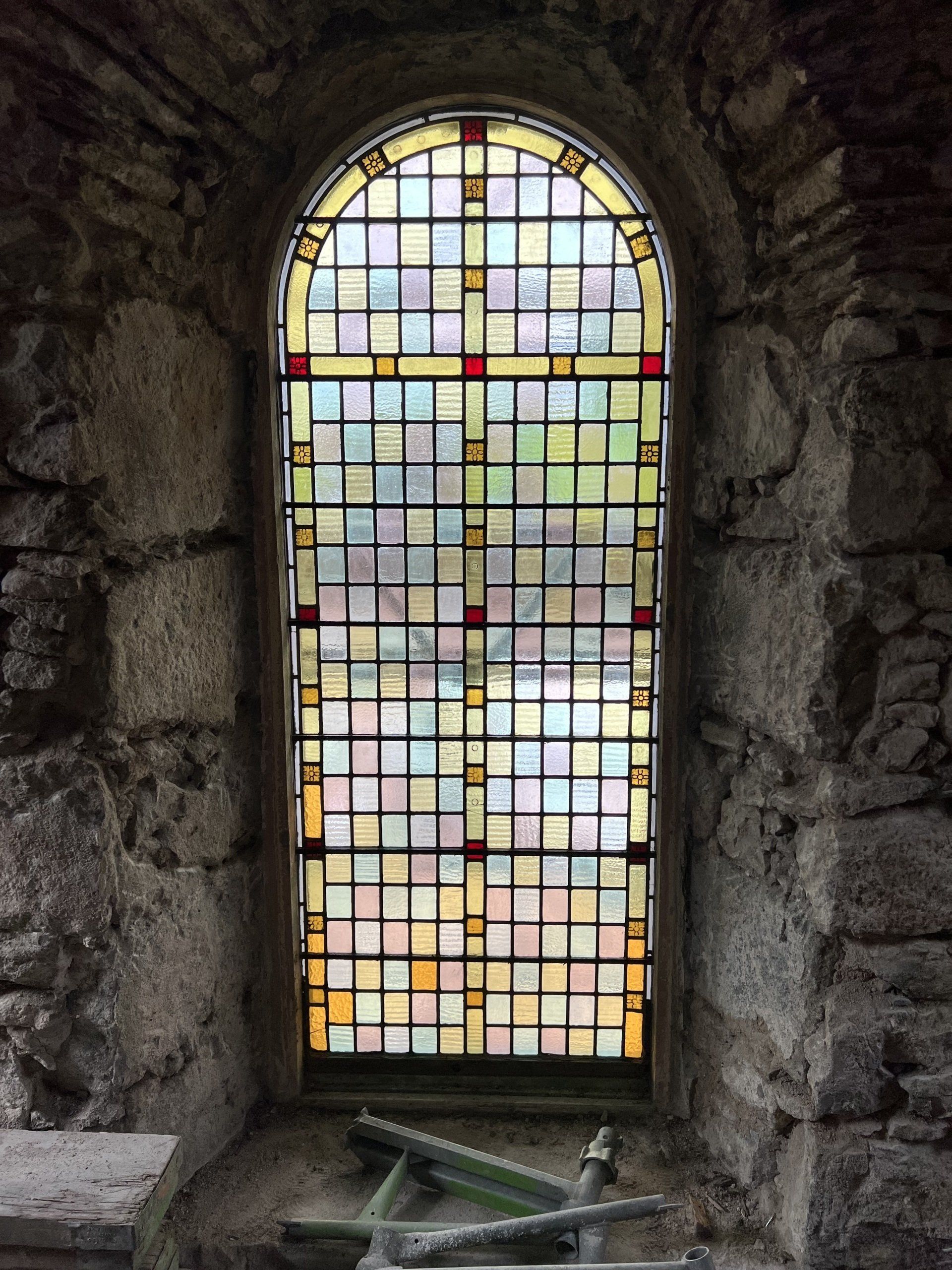 Colorful stained-glass window set in a rough stone wall, with scattered broken pieces at the base.