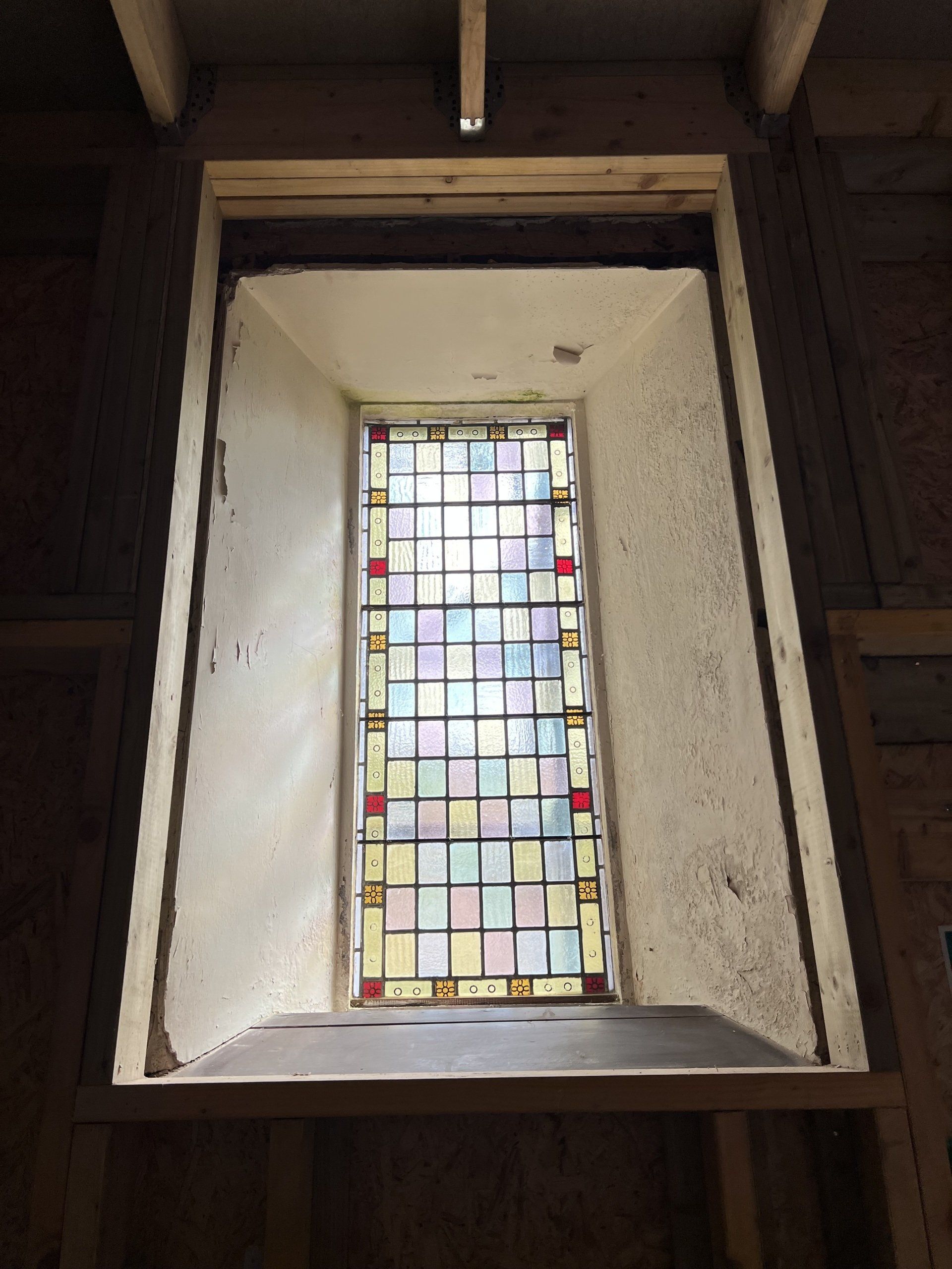 Small stained-glass window set in a wooden wall, framed by a white alcove.