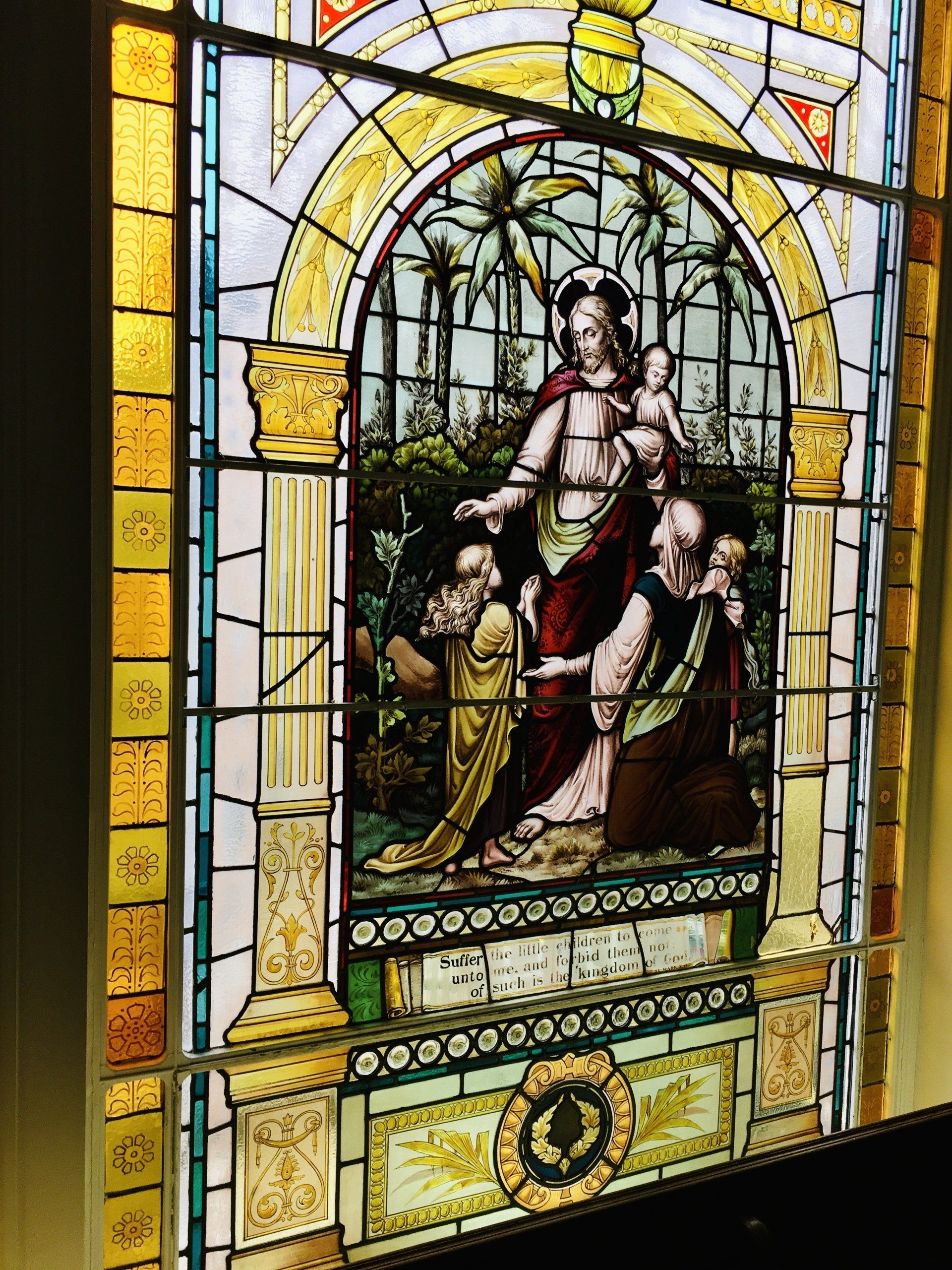 Stained-glass window of a robed figure with a child beneath a palm tree, framed by stone columns.