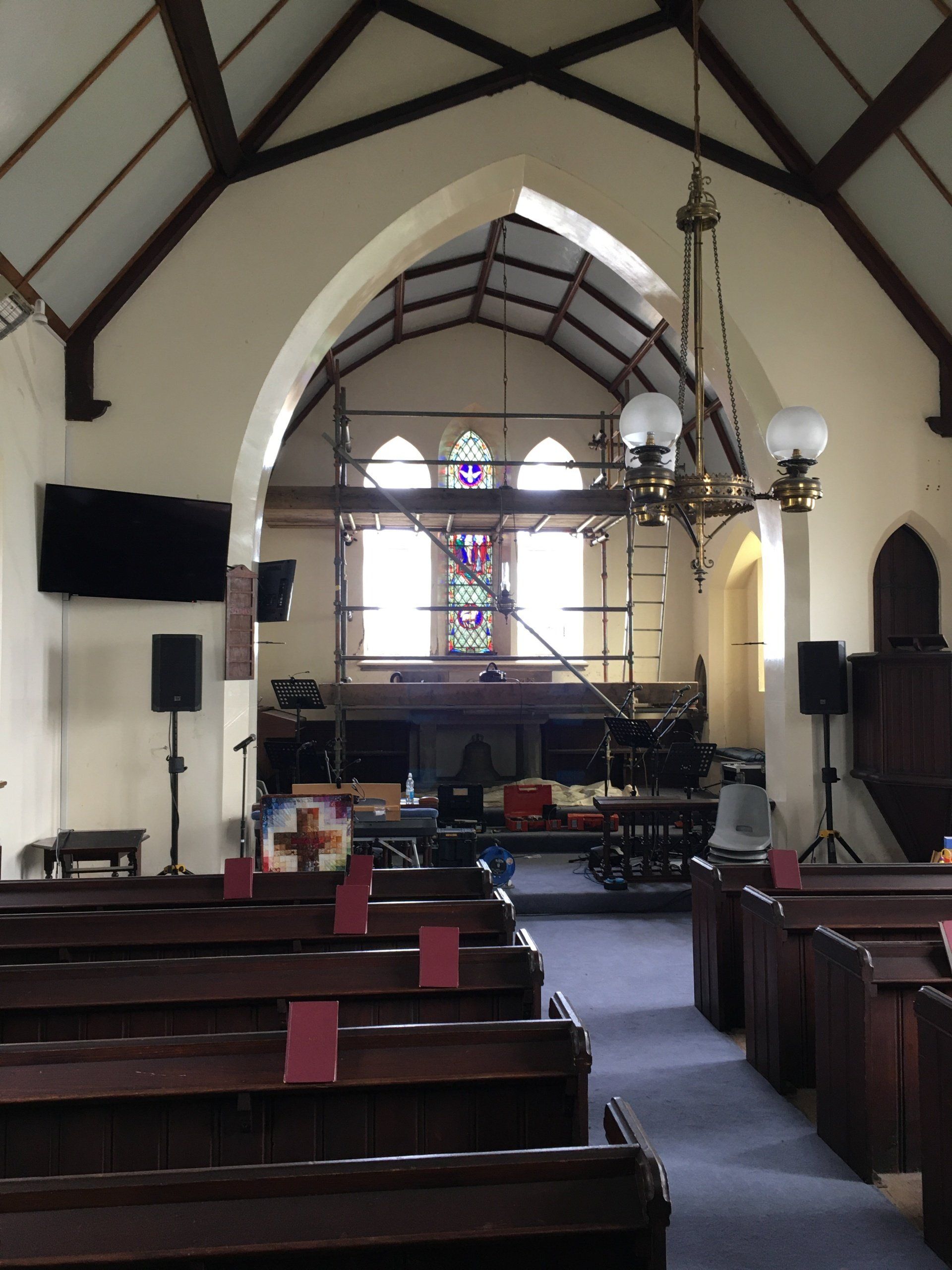 Interior of a small church with wooden pews, stained-glass windows, and a hanging chandelier near the front altar.