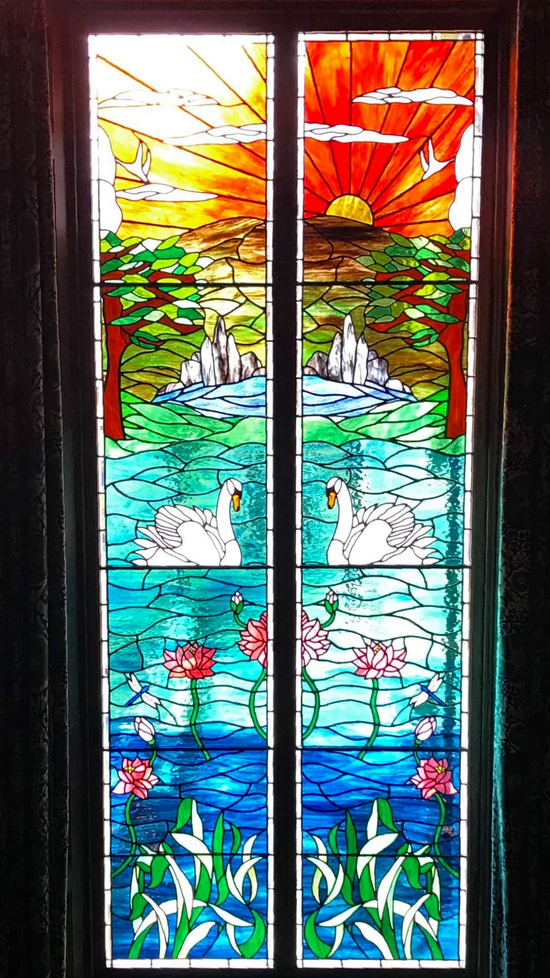 Colorful stained-glass window with a sunrise over water, white birds, flowers, and green hills.