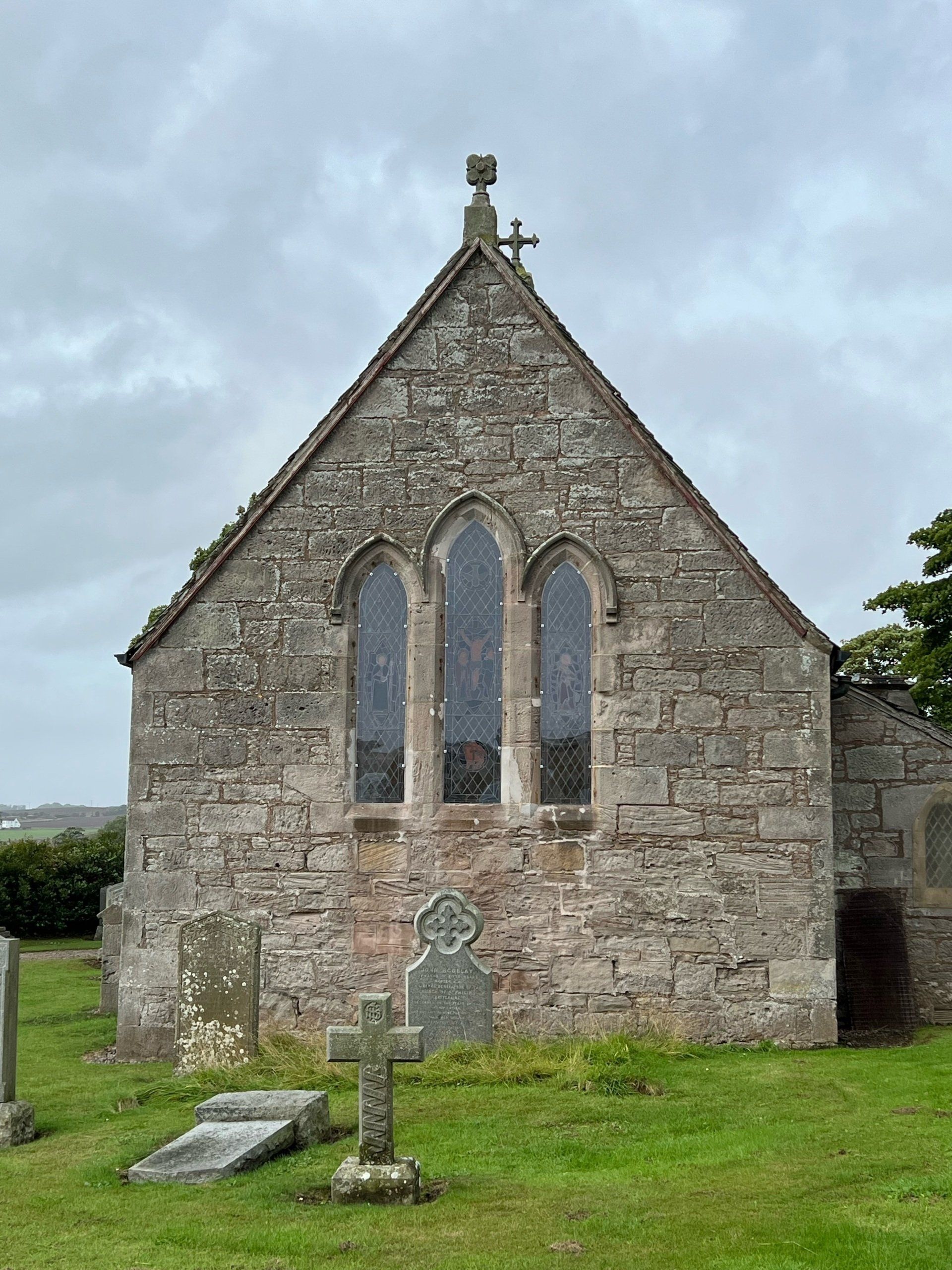 Small stone chapel with arched windows and a cross, set on a grassy field under a cloudy sky