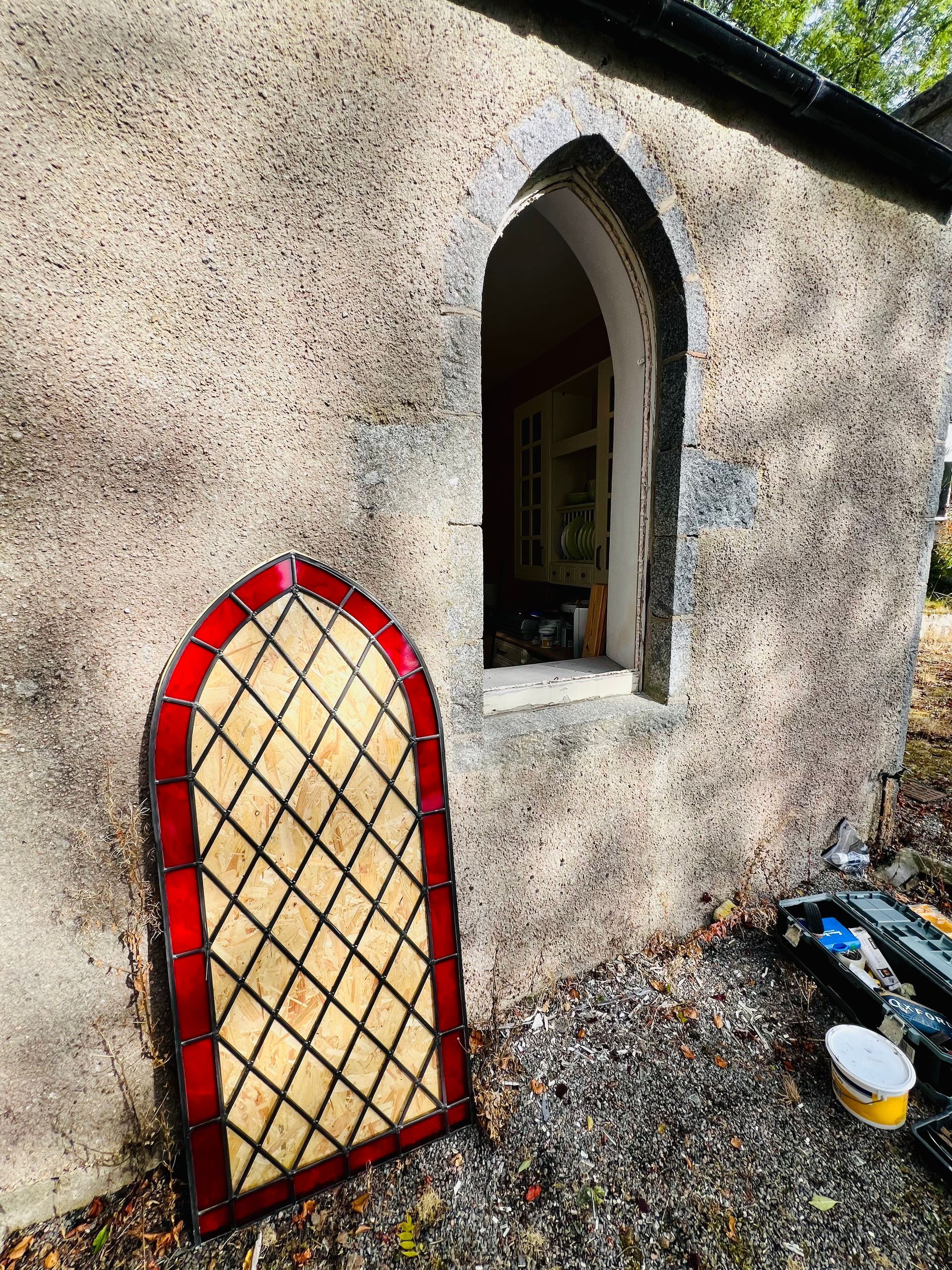 Stone wall with an arched window and a red lattice panel leaning below it.