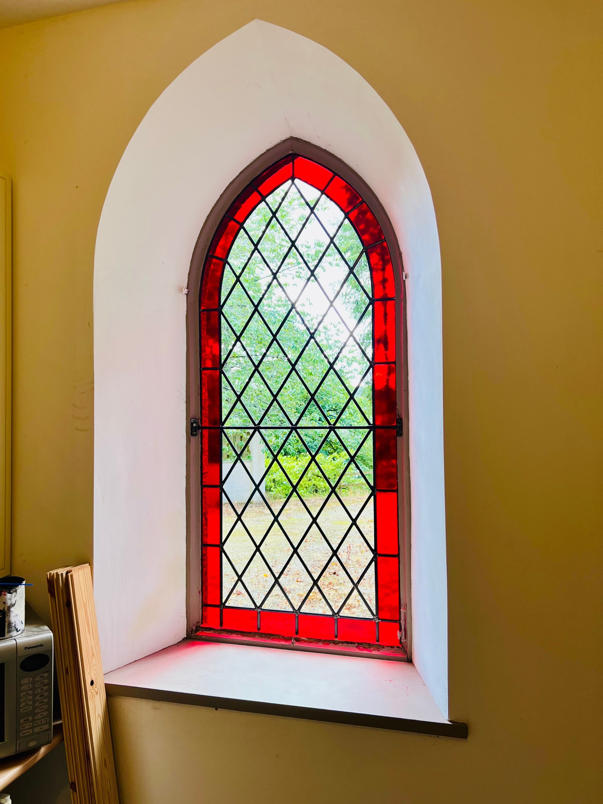Red-framed arched stained-glass window in a cream wall with greenery outside