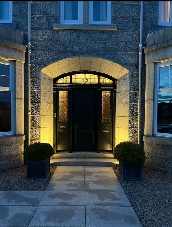 Front entrance of a stone house at dusk, with black double doors, lit archway, and two potted shrubs.