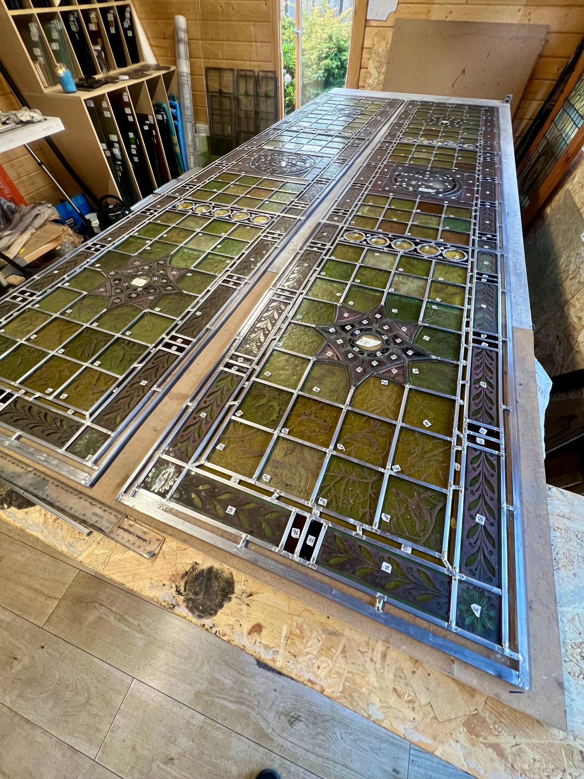 Old stained-glass panel laid flat in a workshop, with green, white, and clear pieces in a wooden frame.