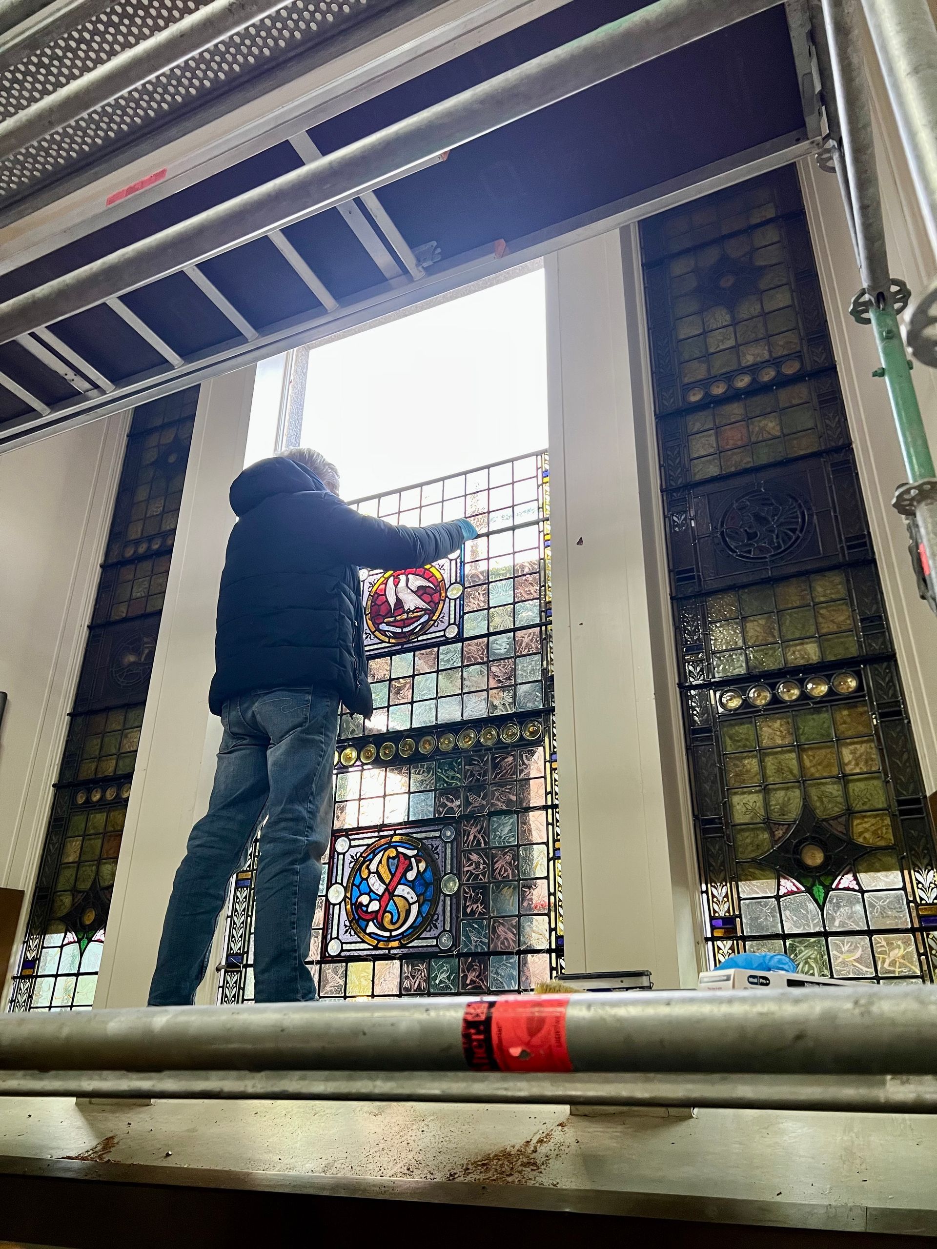 Person standing on a platform adjusting a colorful stained-glass window in a church interior