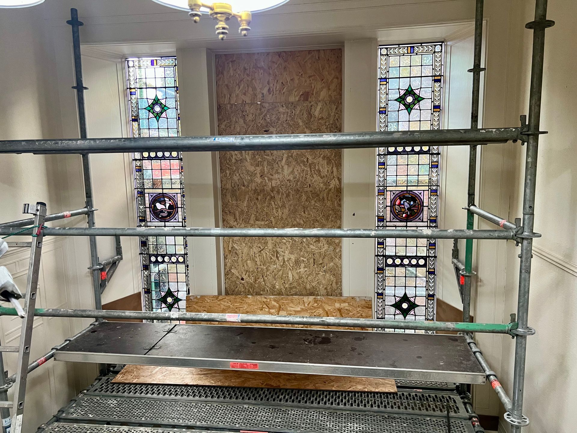 Scaffolding around a boarded-up window area inside a room with stained glass side windows