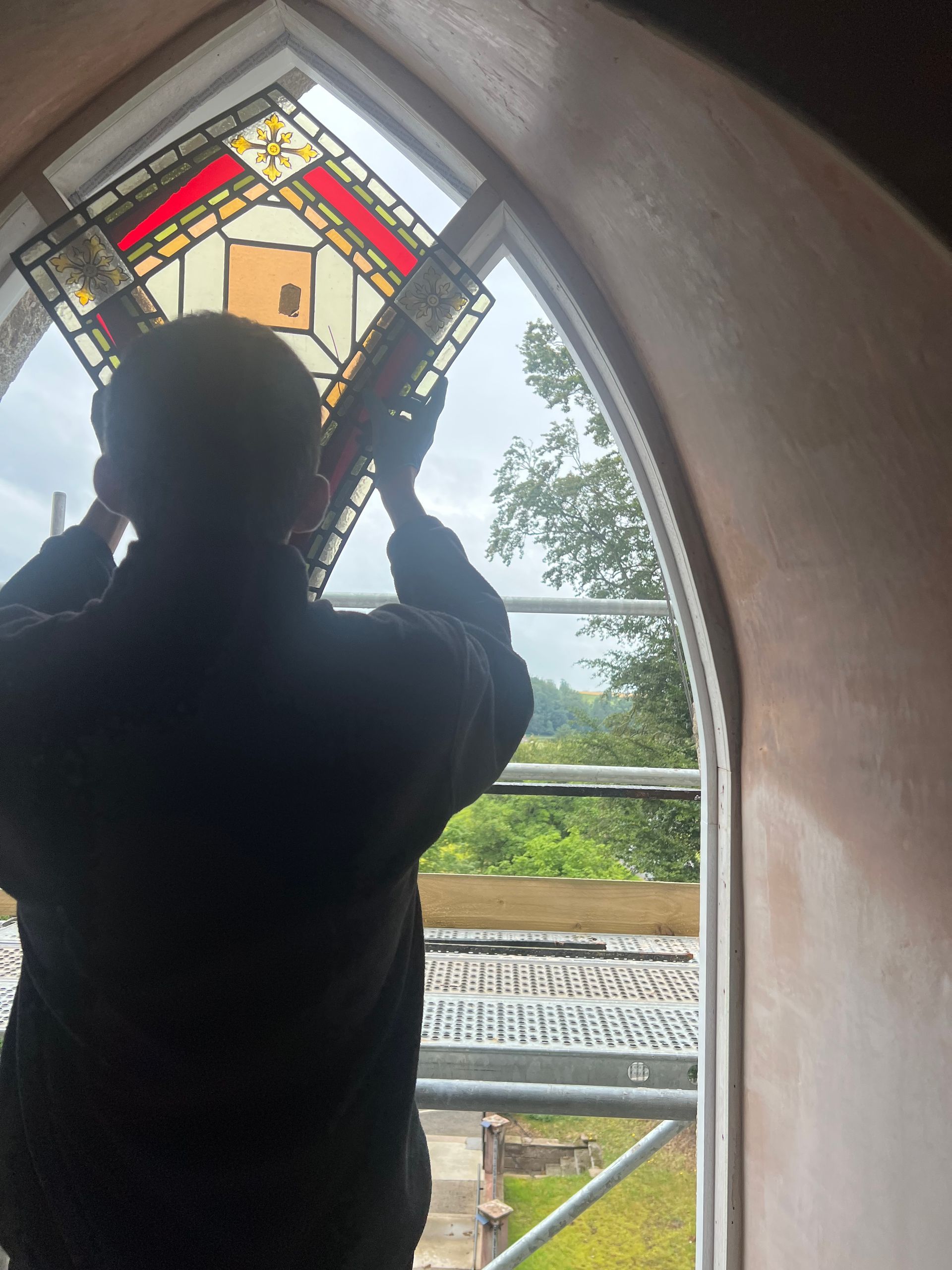 Silhouette of a person reaching up inside a church arch, with stained glass above and greenery outside.