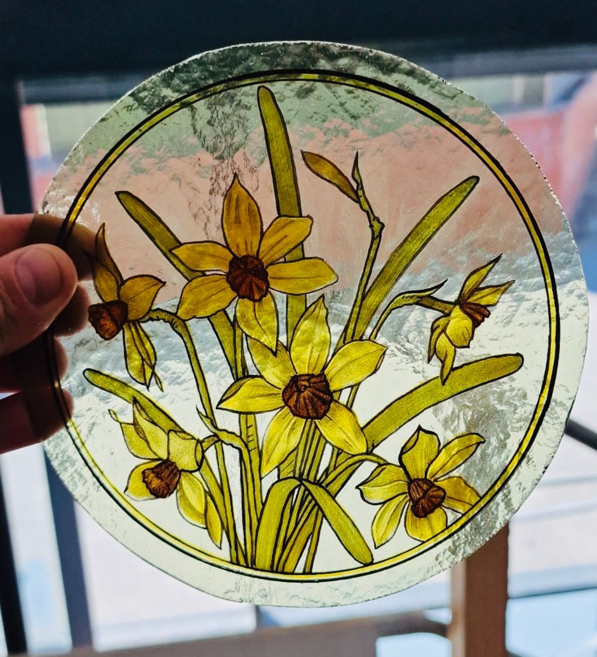 Round stained-glass panel with yellow flowers and green stems held up to a window.