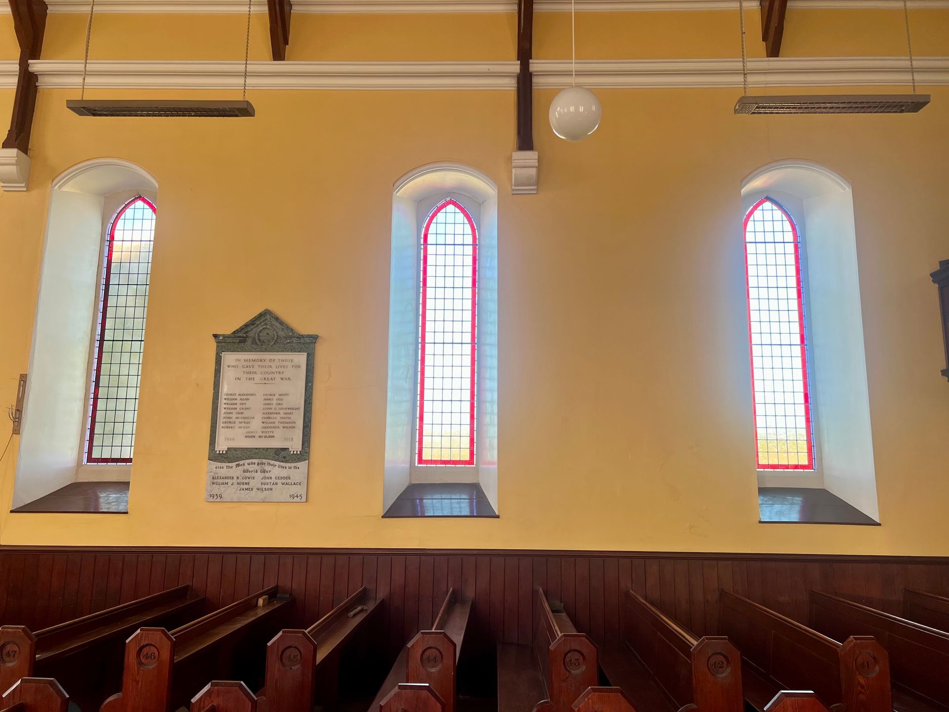 Interior of a church with three stained-glass windows above wooden pews and a memorial plaque on the wall