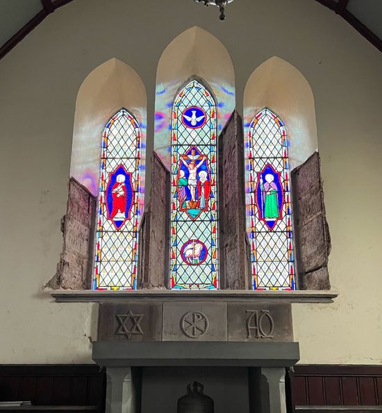 Stained glass church window with three colorful religious figures above a stone mantelpiece.