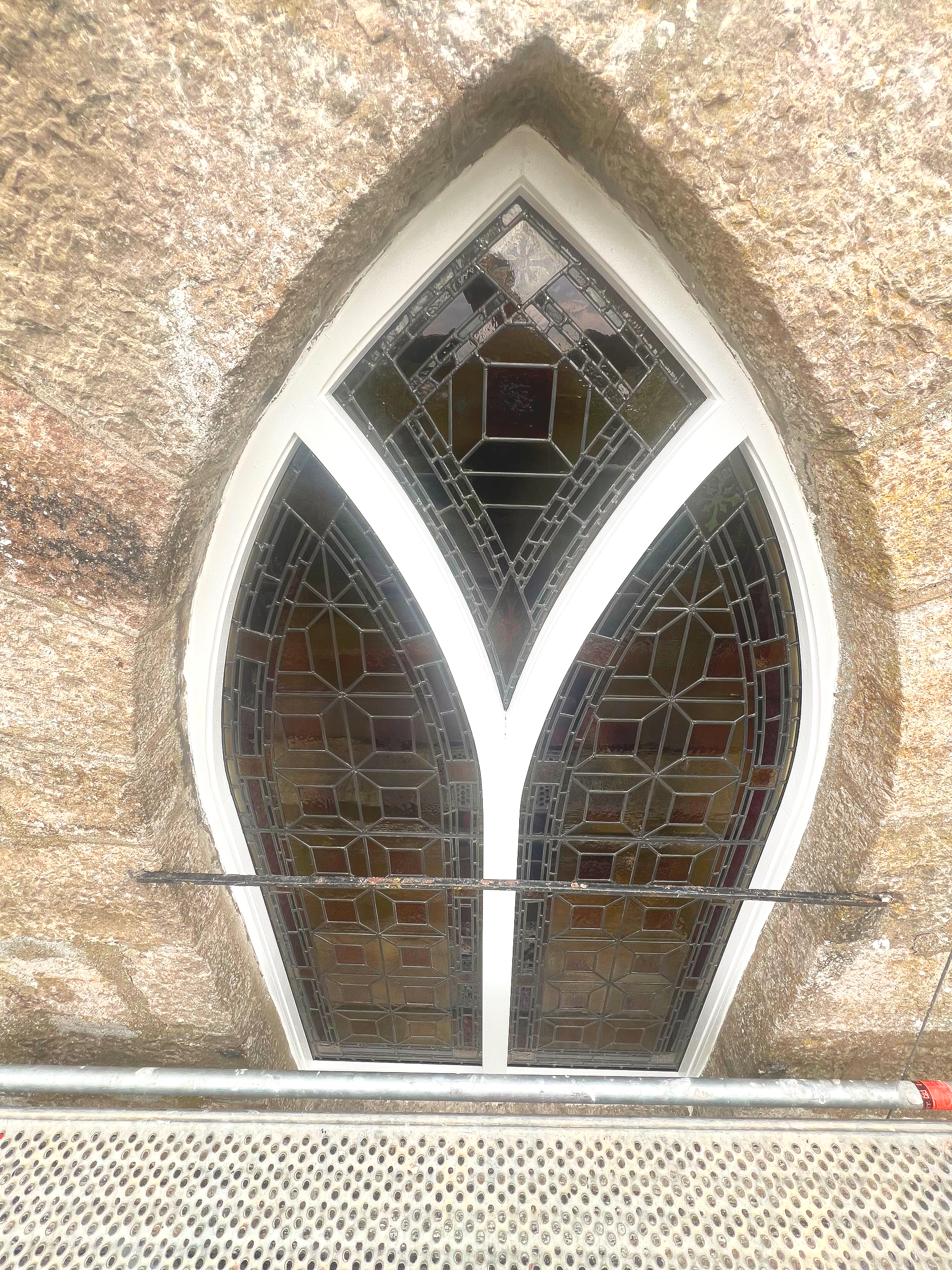Small pointed stained-glass window in a stone wall above a metal grate