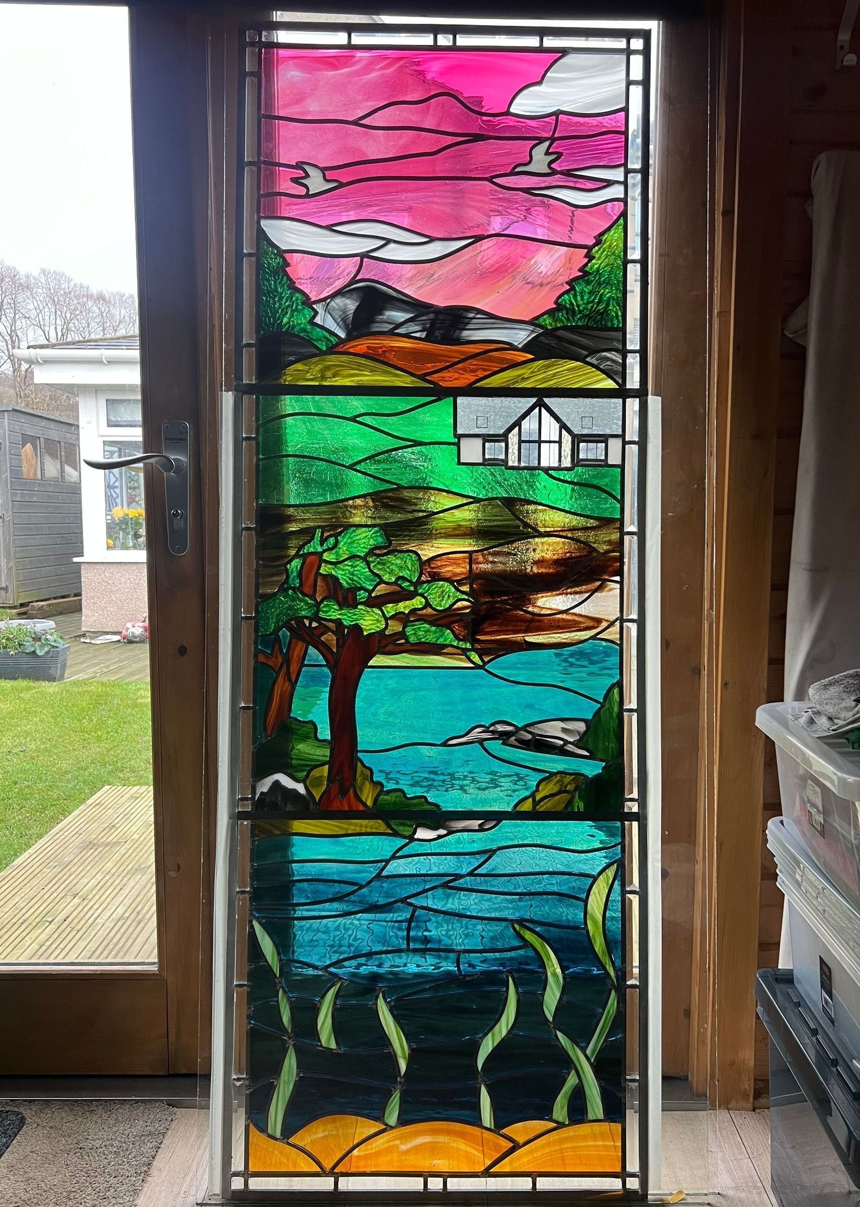 Tall stained-glass panel with pink sky, green tree, house, blue water, and orange flowers in a doorway.